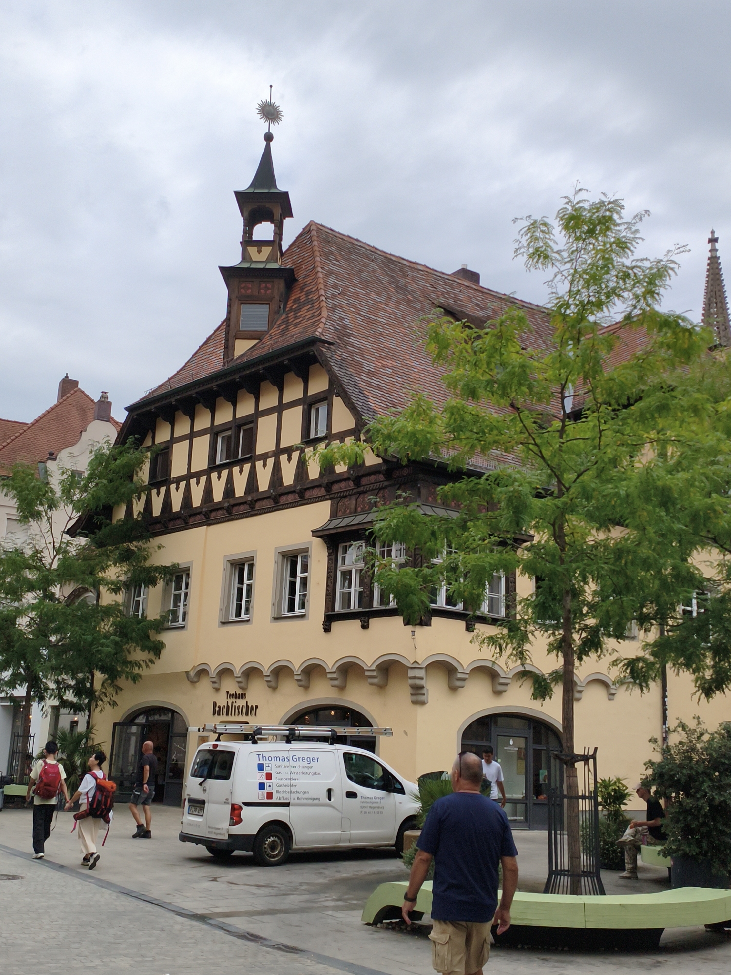 The image captures a street scene in Regensburg, Germany, likely on a cloudy day. In the foreground, a white van with the branding "Thomas Greger" and a list of services is parked. The van has a ladder rack on its roof. To the left of the van, a couple walks away from the camera, both wearing backpacks. The woman is in the foreground, wearing a light-colored outfit and a red backpack, while the man is behind her, wearing a dark shirt, dark pants, and a red backpack.
Behind the van and the couple, a traditional building with a light yellow facade and a red tile roof is visible. The building features intricate timber framing on its upper stories and arched doorways at ground level. One of the doorways has a sign that reads "Teestube Bachfischer". A sun-shaped weather vane adorns the pointed turret on the roof of this building.
To the right of the building, a lush green tree with delicate leaves stands prominently, partially obscuring the facade. Further to the right, a man in a dark shirt and light-colored pants sits on a bench next to dense greenery. Another man in a dark blue t-shirt and khaki shorts is walking towards the bench, away from the camera. The ground is paved with cobblestones or similar material. The overall impression is a charming European street with historic architecture and everyday life unfolding.
