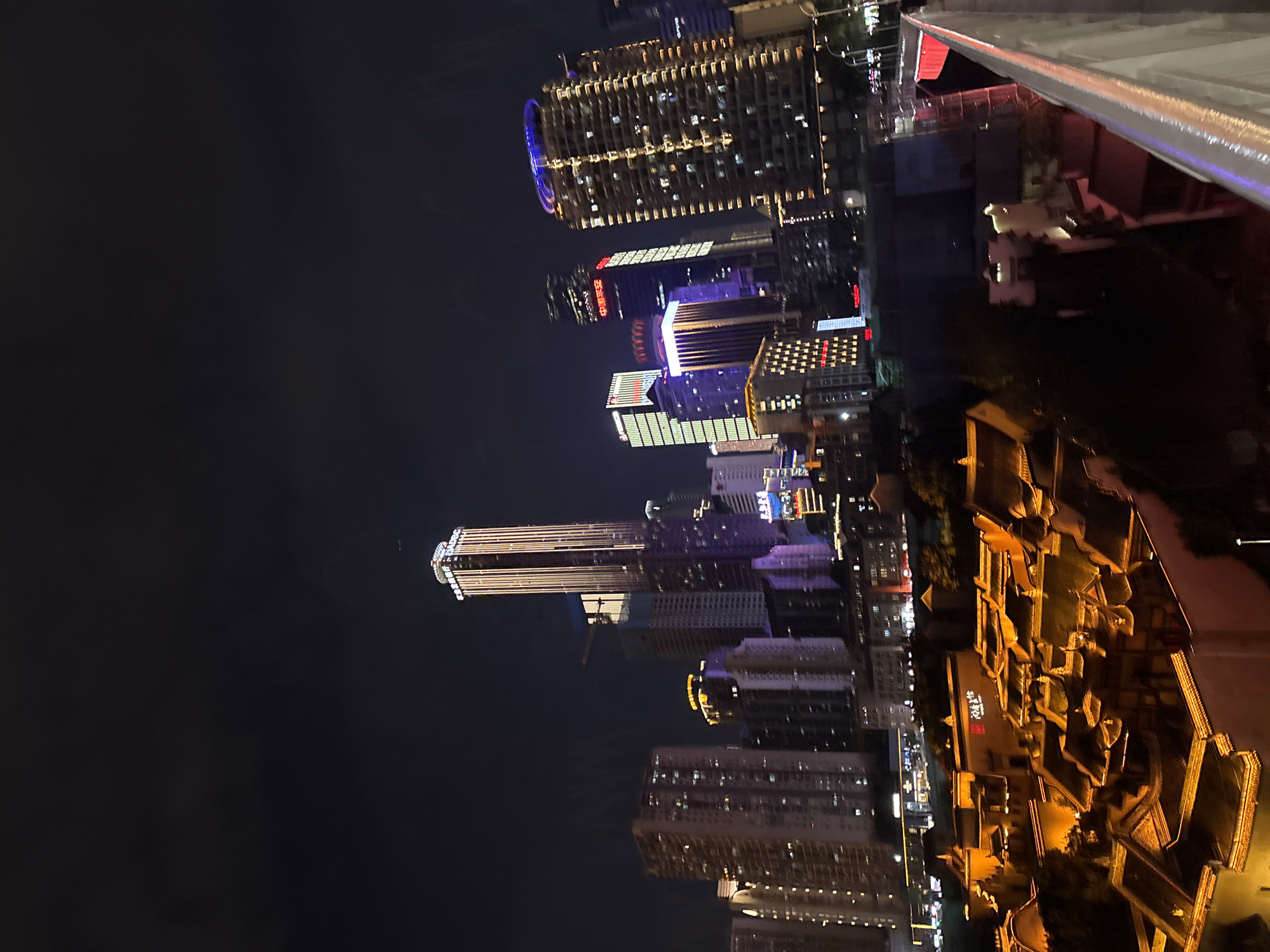 The image displays a nighttime cityscape in Yuzhong District, China, with a focus on illuminated skyscrapers and traditional architecture. The scene is captured from a high vantage point, likely a building or observation deck, as evidenced by the railing in the upper right corner of the image. The skyscrapers are adorned with various lights, including blue, purple, and yellow, creating a vibrant and dynamic urban landscape against the dark sky. One prominent skyscraper features a distinct circular blue light at its apex.
To the right of the modern skyscrapers, there is a section of traditional Chinese architecture with tiered, dark-tiled roofs, illuminated with warm yellow lights. This juxtaposition of old and new architectural styles is a notable feature of the scene. The lighting suggests it is nighttime. The weather appears clear, allowing for a sharp view of the city lights. There are no people visible in the image, and the overall mood is one of urban dynamism and beauty at night.
Visible text appears on one of the traditional buildings, in Chinese characters. The text reads "洪崖洞" (Hongyadong), which is a well-known tourist attraction in Chongqing, located in Yuzhong District. This confirms the location context.