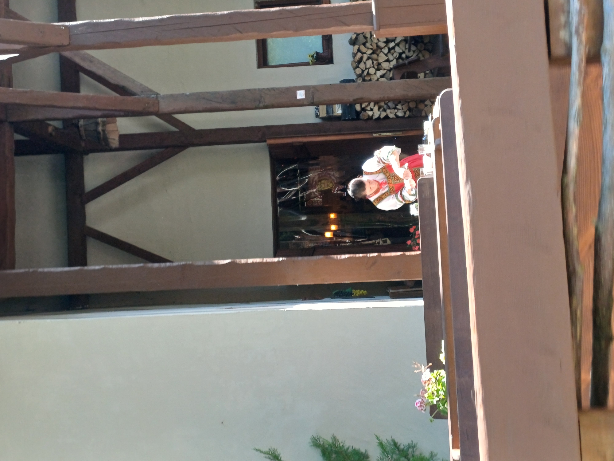 The image shows a woman in traditional clothing at a table in what appears to be an outdoor seating area of a restaurant. The woman is wearing a red and white outfit with embroidery. She is holding a utensil and appears to be preparing food or drink. There are glasses and containers on the table in front of her. In the background, there is a building with wooden beams and a door, as well as a stack of firewood. To the left of the woman, there is a window with a view of potted plants and a green tree. The scene is bathed in sunlight. The location is identified as Estosadok, Russia.