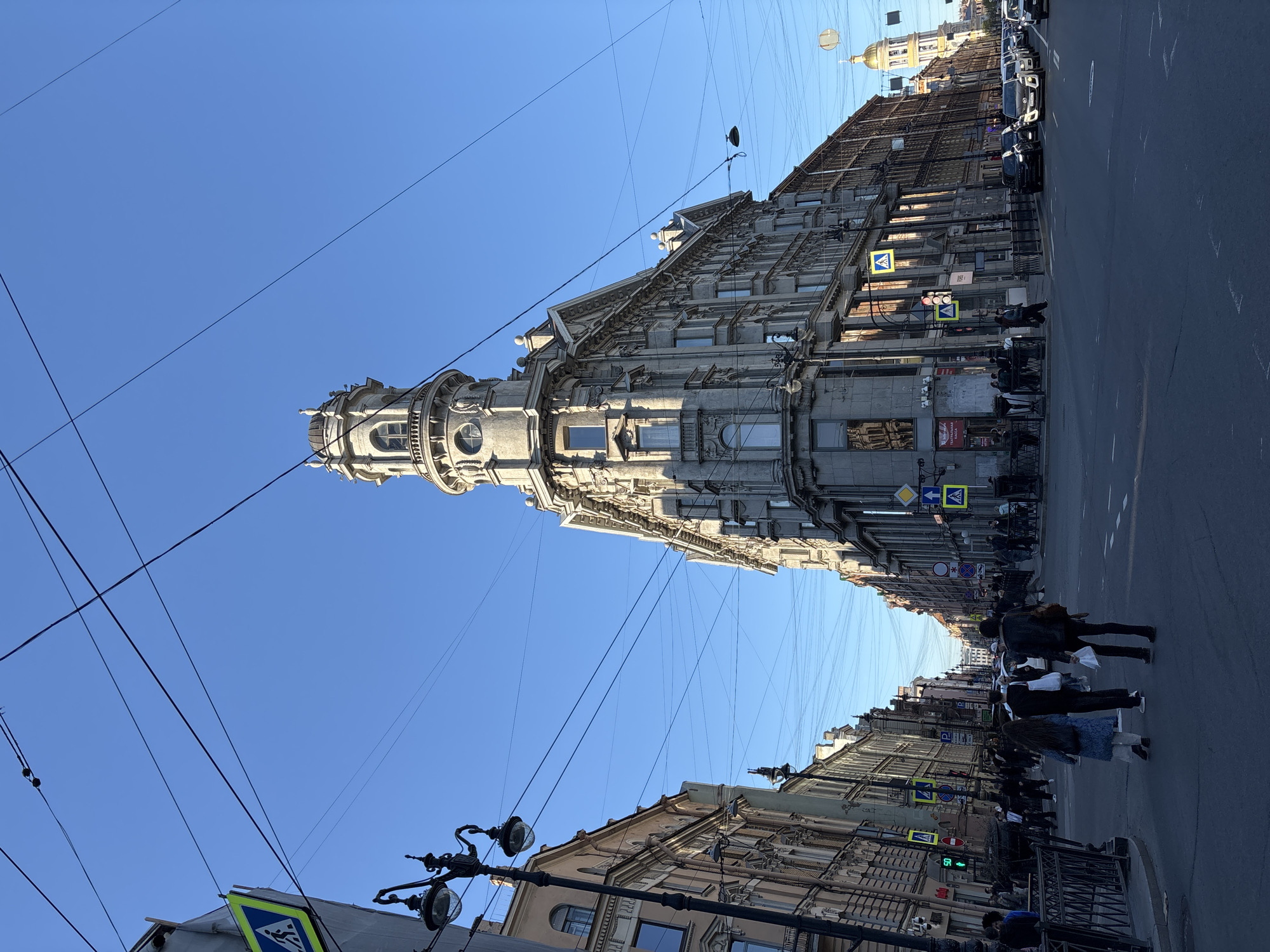 The image captures a street scene in Saint Petersburg, Russia, featuring the landmark known as the Singer House (also known as the House of Books), a notable building in the Art Nouveau style. The building's distinctive tower with a glass dome is prominent against a clear blue sky. Overhead wires stretch across the sky, intersecting the view of the building.
The street is lined with other buildings, typical of the city's architecture, and there is vehicular traffic. People are crossing the street at a designated pedestrian crossing, marked by signs. Streetlights with ornate designs are visible along the sidewalk. Traffic signals are present.