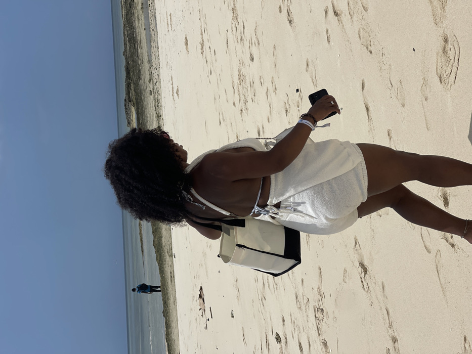 The image displays a bright, sunny beach scene in Zanzibar, Tanzania. A woman, observed from a rear-right perspective, is walking rightward on light-colored sand, leaving footprints. She has dark, curly hair and wears a cream-colored textured two-piece outfit, including a halter top with a tie-back and matching shorts. She carries a cream and black two-tone tote bag over her left shoulder. Her right hand holds a dark-colored smartphone, and she wears a white wristband and a brown beaded bracelet on her right wrist, with a thin chain visible on her left ankle. In the distant background, a person in a blue shirt stands in shallow ocean water, facing away from the camera. The midground water features dark, exposed coral or rock formations. The sky is clear and light blue, indicating fair weather.