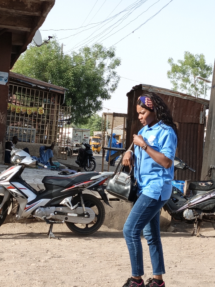 The image depicts an outdoor street scene in Gwagwarwa, Nigeria, under clear daylight.
In the foreground, slightly to the right of the center, a woman is positioned in profile, facing right. She has long, dark, wavy hair partially covered by a patterned headband featuring blue, red, and brown hues. She wears a light blue short-sleeved polo shirt with a white logo on the left chest, dark blue jeans, and dark athletic shoes with pink trim. A dark watch is visible on her left wrist. She holds a black structured handbag in her left hand and appears to be adjusting something in that hand.
To the woman's left, a silver and black motorcycle with red accents is parked on its kickstand, facing right, on the unpaved, dusty ground. Behind this motorcycle, a building with a brown facade and a heavily grated window or entrance is visible. Several yellow rectangular signs or banners are displayed within the grates. Two men are seated on the ground near this building, one dressed in blue attire. A small white satellite dish is mounted on the upper left section of the structure above this building.
Behind the woman and slightly to her right, a weathered structure constructed from rusty corrugated metal sheets is visible. Another parked motorcycle, predominantly dark, is partially visible behind this corrugated structure and to the right of the silver and black motorcycle.
In the midground and background, the scene reveals a dusty street. On the left side of the background, a light-colored truck is parked. Further down the street, a yellow auto-rickshaw (Keke Napep) is visible. A person wearing a blue full-body garment is also present further down the street on the right. Additional buildings with varied construction, including some with corrugated roofs, are visible in the distance. Sparse trees with green foliage are present on both the left and right sides of the background. Multiple thin electrical or communication wires crisscross the clear, light blue sky overhead. The ground throughout the visible area is unpaved and appears to be dirt or dusty earth.