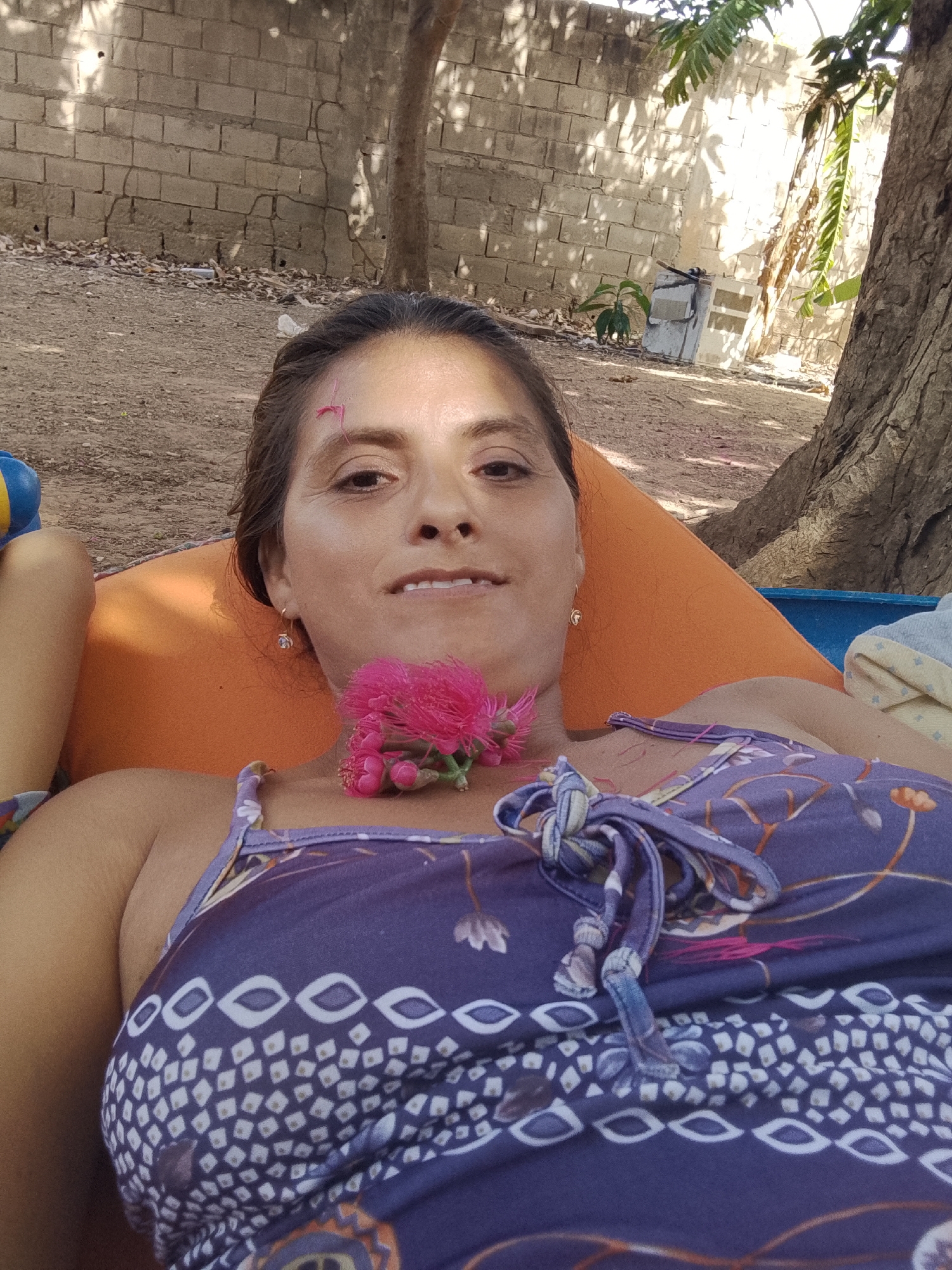 A woman is lying on an orange cushion outdoors, possibly in Altagracia, Venezuela. She is wearing a purple and white patterned top with thin straps. A pink flower is positioned near her neck. She is facing the camera with a slight smile. The background shows a sandy area with trees, a brick wall, and a blue container behind her. A child's limb is visible on the left.