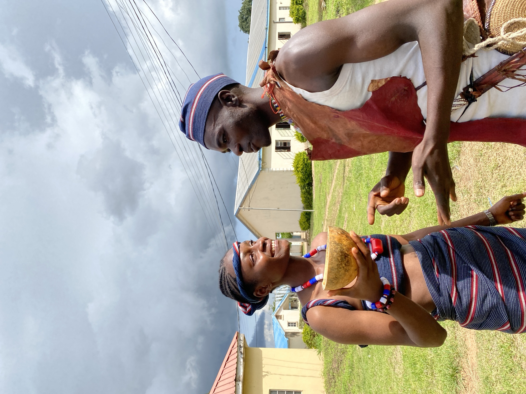 This media file captures a vibrant cultural moment in Wang, Nigeria. Two individuals, a man and a woman, are engaged in conversation outdoors. The man, wearing a white tank top, a traditional leather vest, and a blue and red striped cap, is looking down at the woman. The woman, adorned in a striking striped outfit and a matching headband, smiles warmly as she holds a halved gourd. Her ensemble is further enhanced by colorful beaded necklaces and bracelets.
The scene is set against a backdrop of grassy terrain and several buildings, suggesting a semi-urban or rural environment. The sky is overcast, with dramatic clouds hinting at impending rain. The lighting indicates it is likely daytime, possibly late afternoon given the soft light.
The interaction between the two individuals appears to be friendly and animated, conveyed through their body language and the woman's open smile. The presence of the gourd suggests a possible cultural or ritualistic element to their gathering, perhaps related to traditional crafts or food preparation. The overall impression is one of cultural pride and community interaction within a Nigerian setting. No visible text is present in the image.
