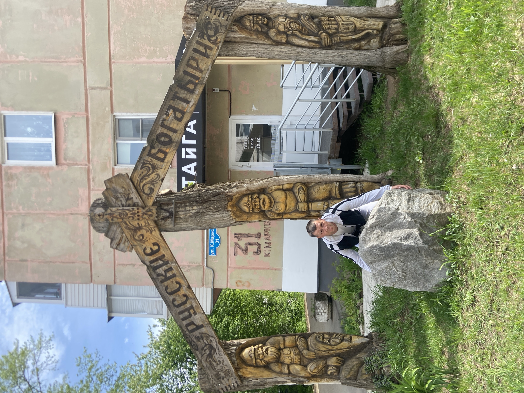 Here's a description of the image:
**General Description:** The photo is taken at ground level, showing a man crouching behind a large gray rock with a wooden structure framing him. The background includes a building and some greenery.
**Location:** The address visible on the building is "ул. К. Маркса, 31" in what I can confidently guess is likely the city of Chaikovsky, Russia.
**People:** A man is crouching behind the rock. He is wearing a white and black jacket.
**Objects:**
* A large, rough gray rock.
* A wooden structure made of what appears to be tree trunks and carved planks. The planks have carved figures and text in Cyrillic.
* A building with a light-colored facade. It has windows and a door. A sign with the text "ТАЙГА" is visible above the door.
**Environment:**
* A patch of green grass surrounds the rock and the wooden structure.
* The sky is visible in the upper left corner.
