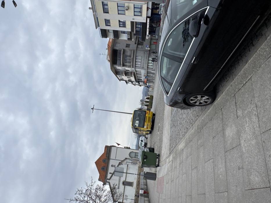 The image depicts an urban street scene in Bragança, Portugal, specifically near the former Bragança train station, identifiable by the "BRAGANCA" lettering on its white facade. The sky is overcast with light grey clouds.
In the immediate foreground on the right, a black four-door sedan, consistent with an Audi A6 model, is parked parallel to a grey stone slab sidewalk. The front of this vehicle is out of frame. The sidewalk extends across the lower left portion of the image.
Adjacent to the sidewalk, the street is paved with dark cobblestones, sloping slightly downwards towards the right. On the left side of the cobbled street, a dark green wheeled waste bin and a dark grey litter bin are positioned near the sidewalk. Behind these bins, a dark-colored car is partially visible.
Further along the cobbled street, a yellow bus is positioned, facing slightly towards the left. Behind the bus, a white commercial van is visible. In the middle-ground, a tall, dark grey street light pole stands.
On the left side of the scene, behind the waste bins, stands a multi-story white building with a prominent reddish-orange tiled roof and several windows. This structure is the former Bragança train station. A tree with bare branches is partially visible on the far left.
On the right side of the street, a sequence of multi-story buildings lines the sidewalk. One building features several balconies with grey railings, and another is predominantly white with blue accents around its windows. Two individuals are observed walking on the sidewalk in front of these buildings, moving away from the viewer. A red postbox is visible on the sidewalk further down on the right. Distant hills are faintly visible under the overcast sky in the background.