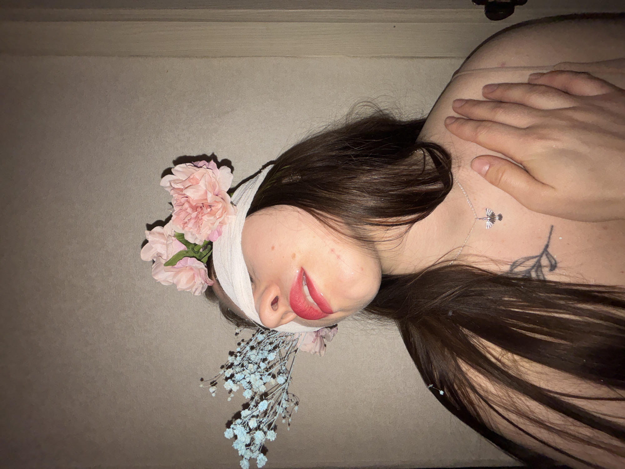 A close-up portrait shows a person, likely female, from the chest up, lying down with their head tilted slightly. They have long, dark brown hair spread around their head and shoulders. A white or off-white band is wrapped around their forehead and the side of their head, serving as a base for floral embellishments.
Tucked into this headpiece on the left side are several soft pink flowers, resembling carnations or peonies. Smaller, delicate light blue baby's breath flowers are arranged near the chin and neck area, blending with the head dressing. The person's eyes are closed, and their lips are parted in a gentle, almost serene expression, adorned with vibrant red lipstick. A few small blemishes are visible on the cheek and chin.
A silver necklace with a distinct pendant, shaped like a stylized bird or fan, rests on the person's chest. On the left collarbone/shoulder, a tattoo depicting a leafy branch design is visible. A hand with natural nails is gently placed on the person's right shoulder/chest. The background is a plain, light brown or beige surface, possibly a wall or floor, with subtle horizontal lines. The lighting is soft, contributing to a calm and artistic atmosphere. No text is visible in the image.