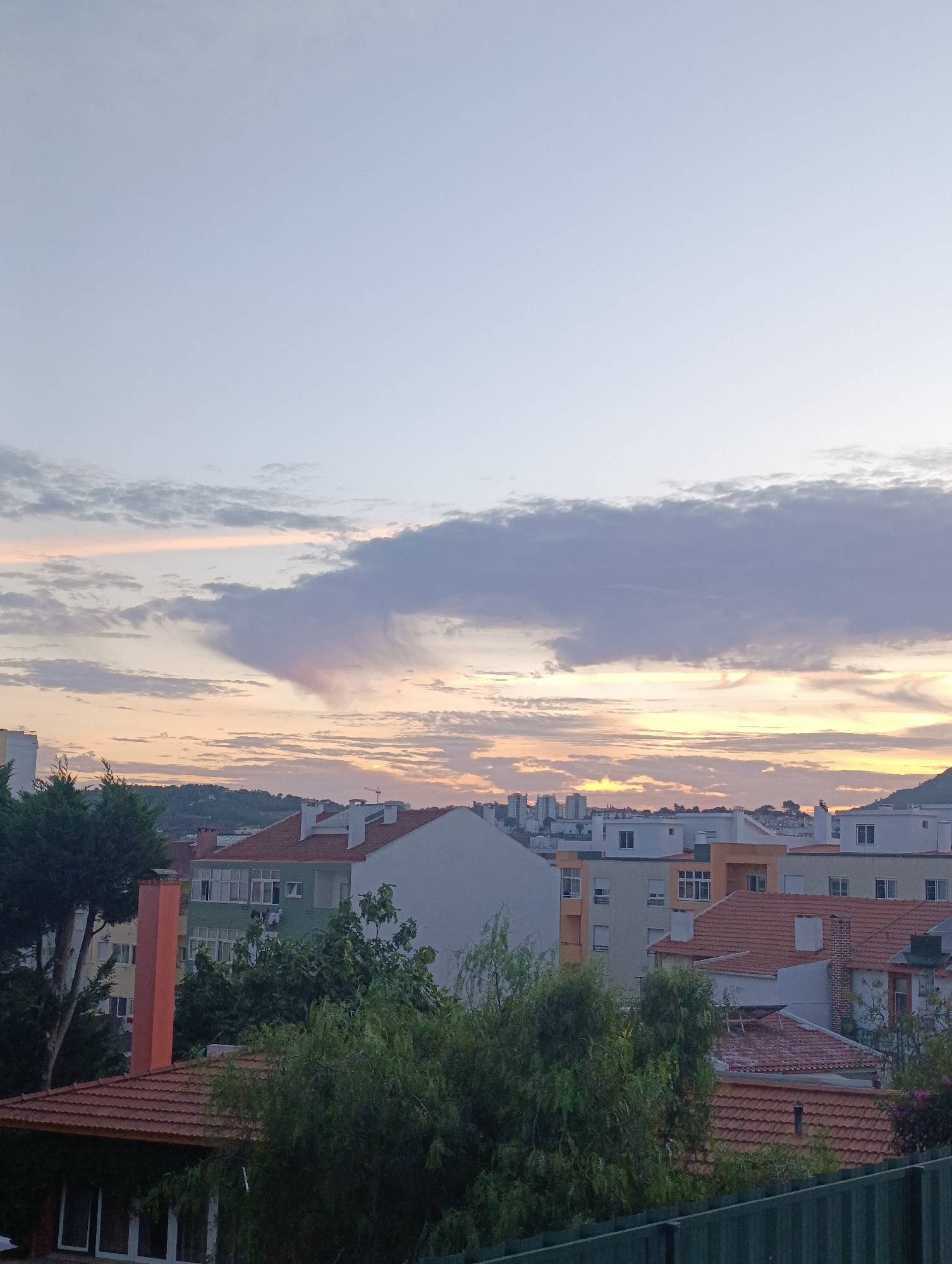The image depicts a view of the city of Setúbal, Portugal, likely taken from an elevated position such as a building or hillside. The time of day appears to be either sunrise or sunset, given the soft, warm hues in the sky and the muted lighting over the cityscape. The sky is a pale blue with scattered clouds that are tinged with orange and pink. In the foreground, there is dense green foliage, including trees and bushes, obscuring some of the lower buildings. Above the foliage, various buildings with terracotta tiled roofs and white or colored facades are visible. Some buildings appear to be residential apartments, while others might be houses. Chimneys are present on several rooftops. In the distance, a more densely packed urban area with taller buildings and a hilly landscape can be seen. The overall impression is a tranquil urban scene at the edge of the day.
