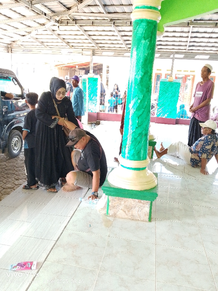 The image shows a scene in front of a building in Anjatan, Indonesia (latitude: -6.419, longitude: 107.979). A woman wearing a black abaya is talking to a man in a black t-shirt and brown shorts while another man sits to the left, observing. Both men are wearing head coverings. The woman's hand is on the man's arm. A green pillar with a white base stands between them, partially obscured by the woman's long black abaya. A second man in a blue shirt sits to the right, with his back to the camera, while a man in a pink shirt sits to the right with a white turban. Both men have their legs outstretched. The background includes a partial view of a white van. There is a large, partially open metal structure with a metal roof overhead. The ground is covered in white square tiles. The scene suggests a public place in Anjatan.