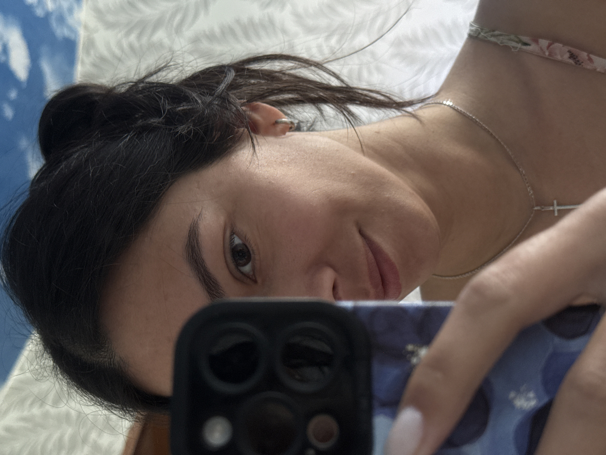 A woman with dark hair and light skin is taking a selfie with a smartphone. The phone is positioned in front of her face, partially obscuring it. She is looking at the camera with a slight smile. The phone has a black camera array. She is wearing a silver necklace with a cross pendant. The background is blurred, but appears to be a light-colored surface with some blue and white patterns. There is no evidence that the photograph was taken in Chaikovsky, Russia.