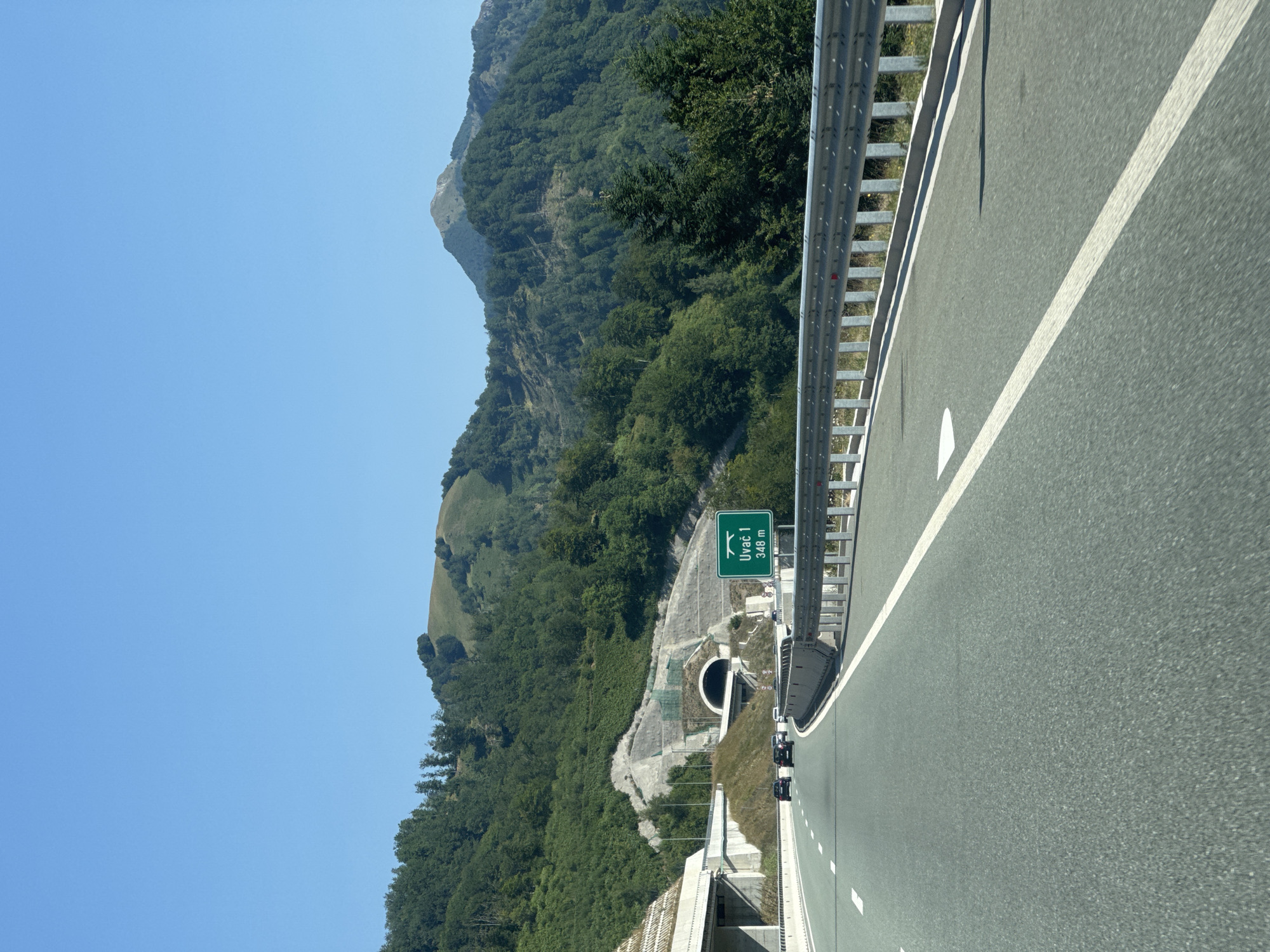 This is an eye-level, point-of-view shot from a vehicle traveling on a multi-lane highway towards a tunnel entrance. The sky is clear and blue. The highway curves slightly to the left and is flanked by a metal guardrail on the right. On the left side of the highway, there is a concrete structure and a grassy embankment leading up to the tunnel. The tunnel entrance is visible in the distance, set into a rocky, forested hillside. A green road sign with white text indicating "Uvač 1 348 m" is positioned on the right side of the highway. Several cars are visible on the highway, moving in the same direction as the viewpoint. The surrounding landscape is mountainous and covered with dense green trees. The image appears to depict a highway in the Tuzi Ljevorječke area of Montenegro, given the road sign.