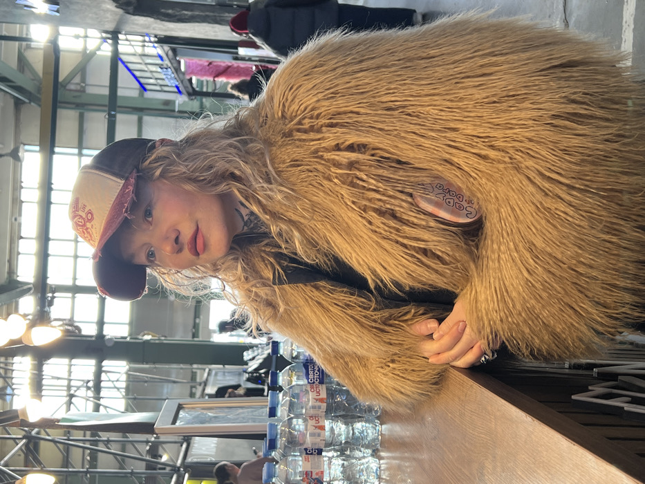 A fair-skinned female with wavy blonde hair, wearing a distressed baseball cap with a cream front, red brim, and dark mesh back, is captured from the chest up, positioned at a wooden counter. She is adorned in a shaggy, light brown faux fur coat, under which a dark garment is visible. Tattoos are present on her neck. Her hands are clasped on the counter's surface, and a pin featuring "Lady S(a)ra" text is affixed to her right lapel. Her gaze is directed towards the upper right of the frame.
The background reveals a large, brightly lit indoor space with high ceilings, exposed metal beams, and large windows allowing natural light. Multiple pendant lights with exposed bulbs illuminate the area. On the counter to the immediate left of the subject, several 0.5-liter plastic bottles of "Святой Источник" (Svyatoy Istochnik) mineral water are arranged. Blurred figures of other individuals are visible in the mid-ground and background, moving or standing, suggesting a public, active environment. One individual in the background wears a red beanie and a dark puffer jacket. The architectural elements and overall setting indicate a repurposed industrial building, consistent with the Vasileostrovsky Market in Saint Petersburg, Russia, known for its food hall and market stalls.