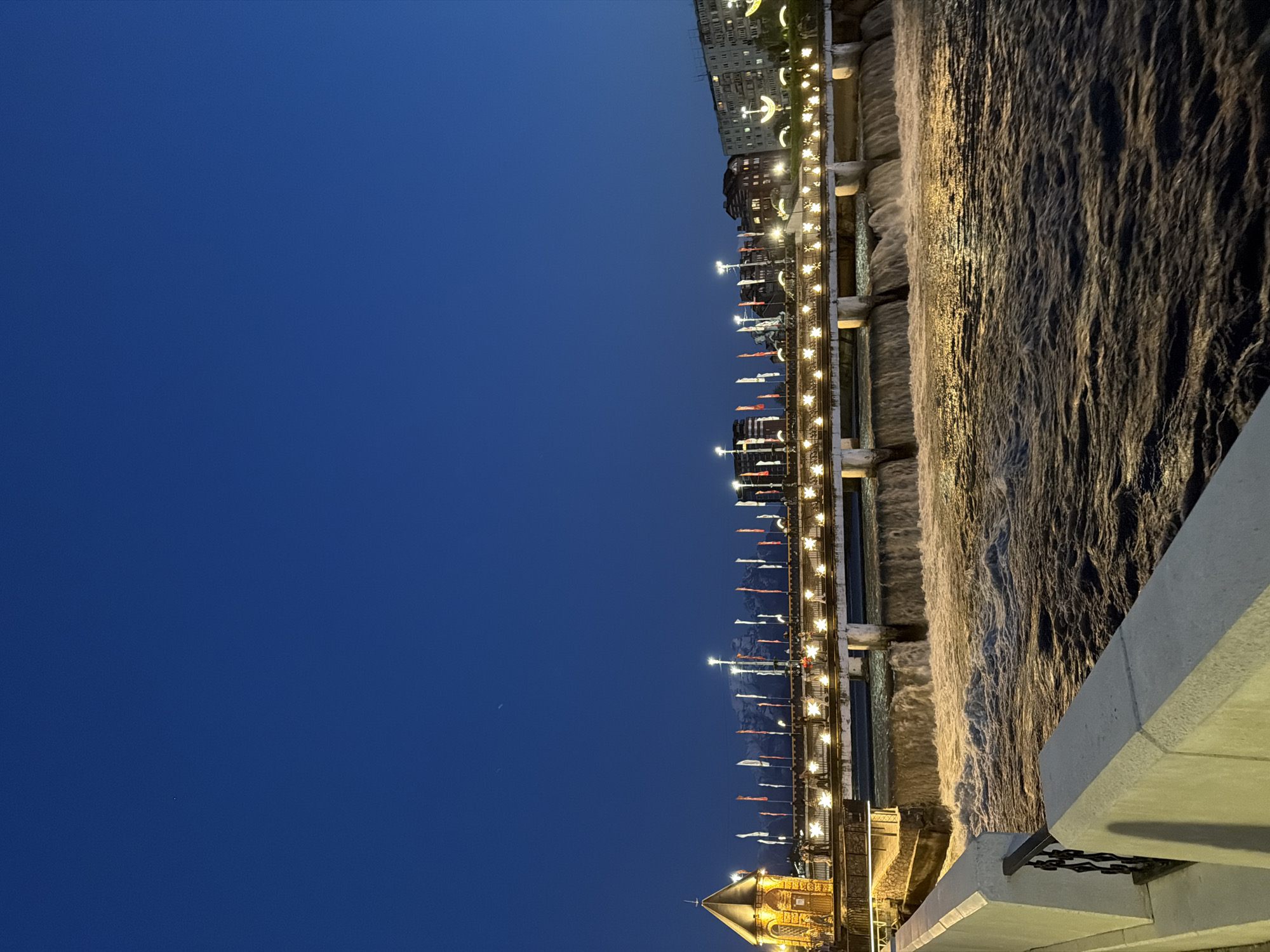 The image presents a nighttime scene in Chermen, Russia, featuring a river flowing beside an illuminated bridge. The sky above is a deep blue. The river's surface reflects the lights from the bridge, creating a shimmering effect. The bridge is lined with lampposts and what appear to be banners or flags. Alongside the river is a concrete barrier. Buildings can be seen further along.