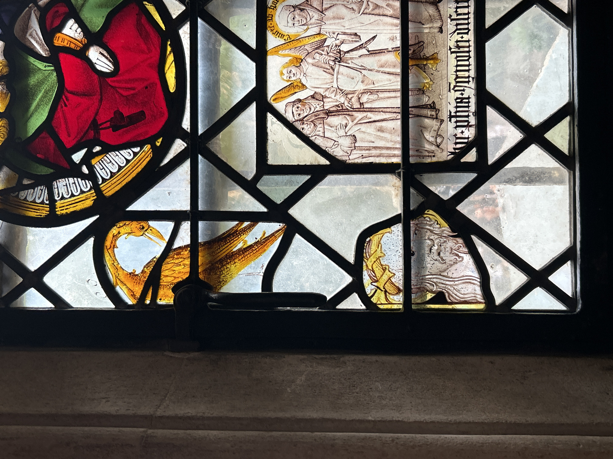 The image shows a close-up view of a stained-glass window, likely in a historic building in Cambridge, United Kingdom. The window is divided into geometric panes by leaded lines.
In the upper portion of the window, a stylized golden bird with prominent feathers and a long neck is depicted. Adjacent to this, a figure dressed in a vibrant red robe with green accents is visible. Their hands are positioned near the robe, with one hand appearing to hold something.
Below this, a scene with multiple figures in robes is rendered. Two figures on the left appear to be in conversation or in contemplation, while a winged figure, possibly an angel, stands between them and another robed figure on the right. The winged figure holds a staff or rod. Inscribed text is present below these figures.
In the bottom left of the window, a partial depiction of a bearded face with long hair is visible, with a golden element above it. The background of the window is clear glass, allowing views of the outdoors, which appear to be greenery. The surrounding stonework of the building is visible on the left side of the image.