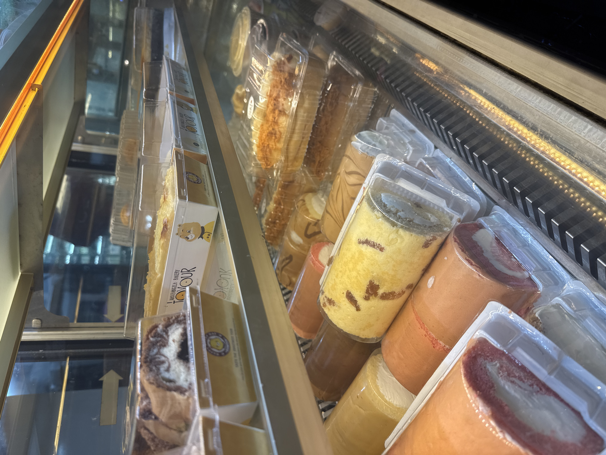 The image presents a detailed view of a refrigerated glass display case filled with packaged baked goods, located in Bac Nha Trang Ward, Vietnam.
**Top Shelf:**
The upper left section of the display case contains multiple rectangular clear plastic containers, each housing a segment of a swirled chocolate and light-colored cake roll with a visible cream filling. These containers are placed on white cardboard trays, one of which is clearly branded "MATOANGELA BAKERY TOYOUR" alongside a cartoon illustration of a brown bear. To the right, additional clear plastic containers hold flat, light-colored baked items, likely tarts or square cakes, also resting on similarly branded "TOYOUR" trays. The glass panel above reflects two white arrow signs pointing horizontally to the right, indicating reflections of the surrounding environment. An overhead orange-yellow light strip illuminates the contents of the display case.
**Middle Shelf:**
The middle shelf features a front row of approximately six individually plastic-wrapped Swiss roll type cakes. Visible colors include beige, brown, red, orange, and yellow, with some rolls displaying distinct dark flecks. Each roll contains a prominent white or light-colored cream filling. Behind this front row, several elongated pastries are visible within clear rectangular plastic containers. These pastries are light brown in color and appear topped with shredded cheese or a similar granular ingredient.
**Bottom Shelf:**
The bottom shelf contains further packaged baked goods, including more Swiss rolls similar to those on the middle shelf, though their details are partially obscured. The base of the display case is composed of a silver-colored, horizontally ribbed metal grate, featuring numerous small, embedded orange-yellow light sources that create a patterned illumination across its surface.
No individuals are visible within the frame. The scene is static, showcasing the bakery products in a commercial setting.