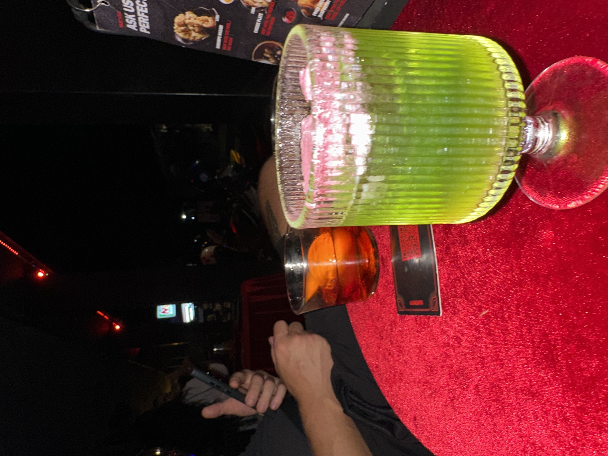 The image shows a low-light indoor scene, presumably in a bar or restaurant in Canggu, Indonesia, as evidenced by the menu item descriptions on the table. A person is holding a phone in their left hand. On the red velvet tablecloth are two cocktails: one green, tall, and ornate with pink elements and a stem, and another amber-colored drink in a smaller glass. There is a menu on the table. In the background are lights suggesting a cityscape or busy street, with the logo of 7-Eleven visible.