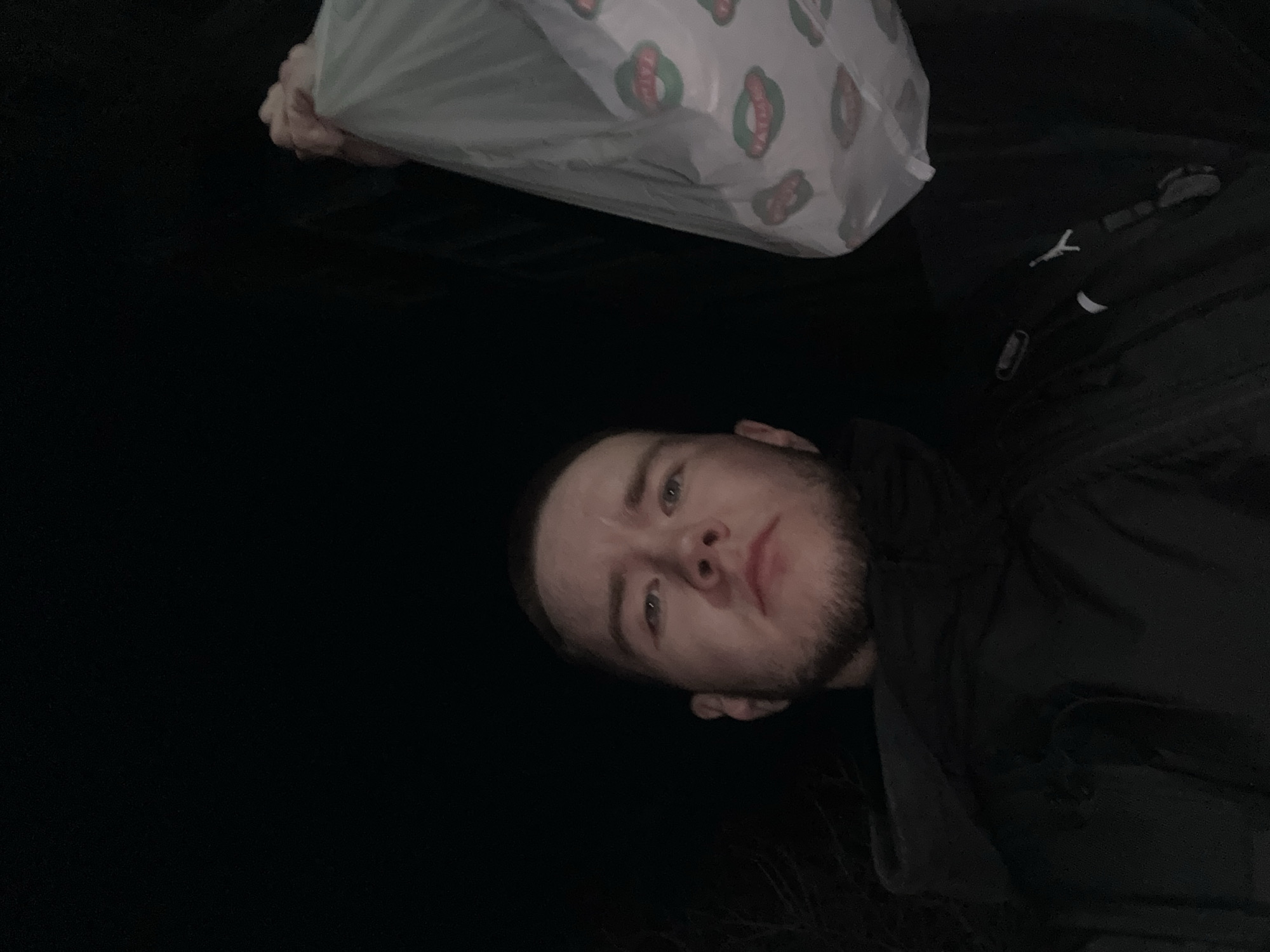 The image is a selfie portrait of a young man, rotated 90 degrees counter-clockwise, appearing to be taken at night in Kazan, Russia, given the extremely dark background. The man, with light-colored eyes and a short beard, looks directly at the camera with a neutral expression. He is wearing a dark-colored jacket or hoodie.
In the upper portion of the image, the man holds a white, semi-translucent plastic bag with his left hand. The bag is patterned with repeating oval-shaped logos, each featuring a green border, a red interior, and a stylized depiction of what appears to be a hot dog or sausage in a bun. This suggests the bag is from a fast-food establishment or a store selling such items. The background is almost entirely black, offering no discernible details of the location or surroundings, emphasizing the nighttime setting.