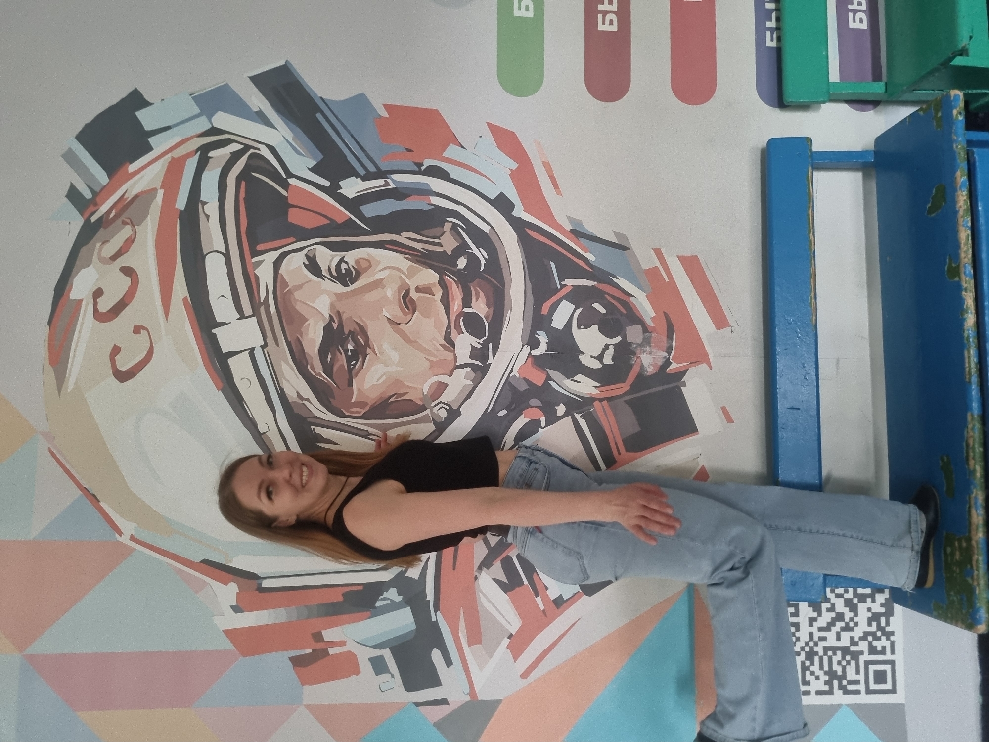 A young woman is lying in front of a mural depicting Yuri Gagarin in Chaikovsky, Russia. She is wearing a black top and blue jeans, and she is smiling. The mural is colorful, with geometric shapes in the background. To the right of the mural is some blue furniture. A QR code is visible on the floor near the woman's feet.
