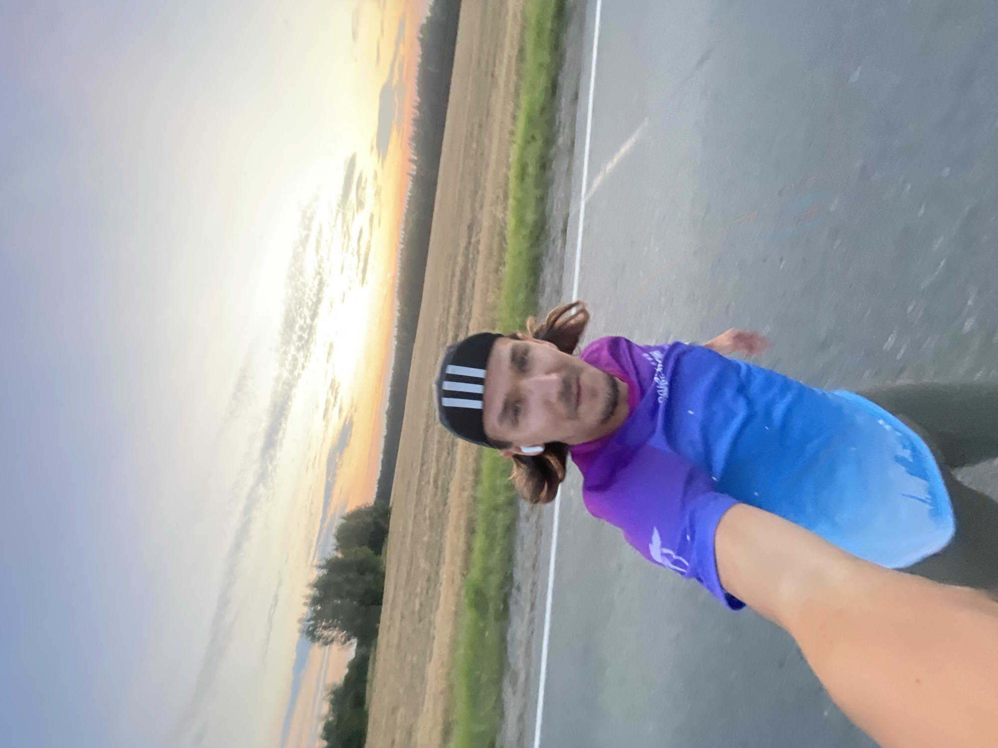 A selfie-style shot shows a Caucasian man with brown hair, a beard, and a black and white Adidas baseball cap with three white stripes, wearing a purple and blue ombre t-shirt and dark leggings, on an asphalt road, possibly a highway, at dusk. The man is wearing white wireless earbuds. He is looking at the camera with a slight smile. In the background, a field of harvested crops stretches to the horizon, bordered by a line of trees. The sky is a gradient of pale blue and orange, with wispy clouds. The image is taken from a low angle, implying the man is running or cycling. The general location is in or around Ol'khovo, Russia, indicated by the rural setting and vegetation.