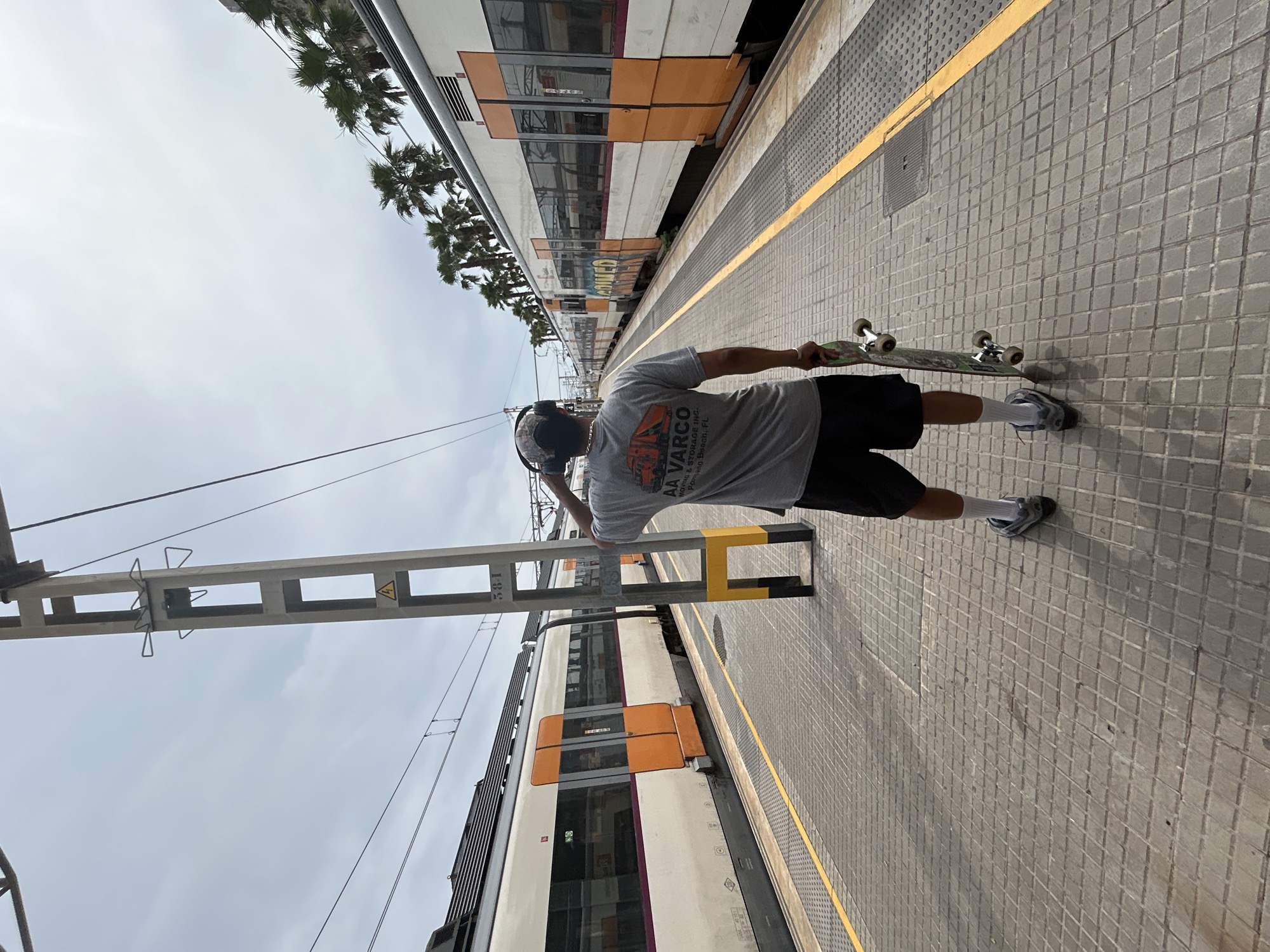 This image captures an outdoor train station platform in Mataró, Spain, under an overcast sky. A male individual is centrally positioned, seen from behind, standing on the grey paved platform. He wears a grey short-sleeve t-shirt with a red and black "AA VARCO Moving & Storage Inc. Orlando Beach, FL" logo, black shorts, white crew socks, grey athletic shoes, a camouflage baseball cap, and black over-ear headphones. His right hand is raised towards his right ear. In his left hand, he holds a skateboard with a green underside featuring graphics and visible wheels.
Two multi-car passenger trains, predominantly white with orange doors and accents, are stationary on tracks flanking the platform. The train on the right exhibits faded yellow graffiti near its forward section. A concrete utility pole with a black and yellow painted base and a yellow triangular electrical hazard symbol is positioned to the left of the individual, partially obscuring the train on the left. The pole also has a label "5381". Overhead electrical lines (catenary system) span across the tracks. A yellow tactile warning strip runs along the edge of the platform. Beyond the train on the right, several palm trees are visible.