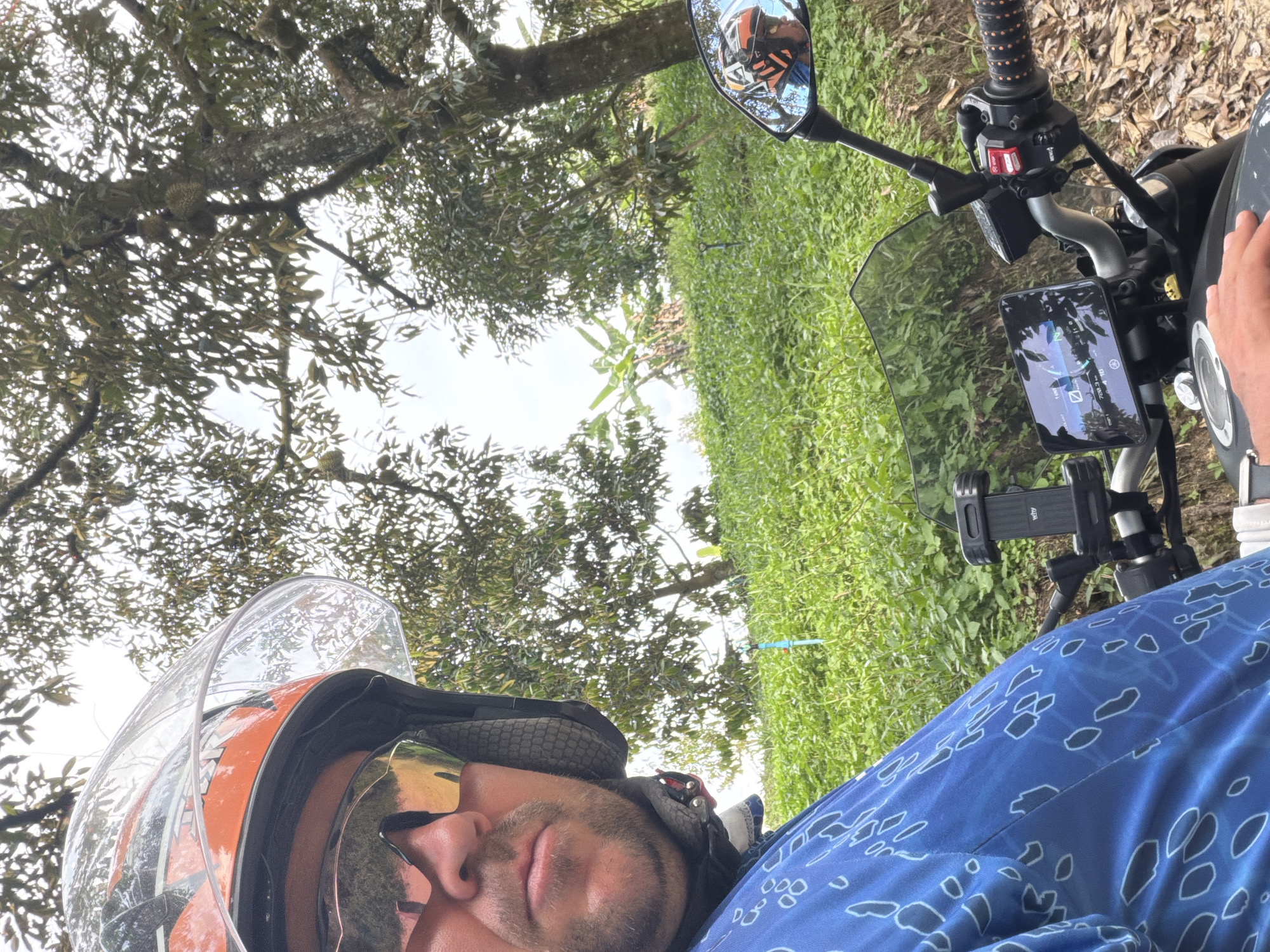 A man wearing an orange and black helmet and sunglasses is riding a motorcycle through a field in Baan Tai, Thailand. The motorcycle has a windshield, a digital display showing speed and distance, and a phone mount. The man is wearing a blue shirt with a white abstract pattern. In the background, there are durian trees and lush green vegetation. The sky is overcast.