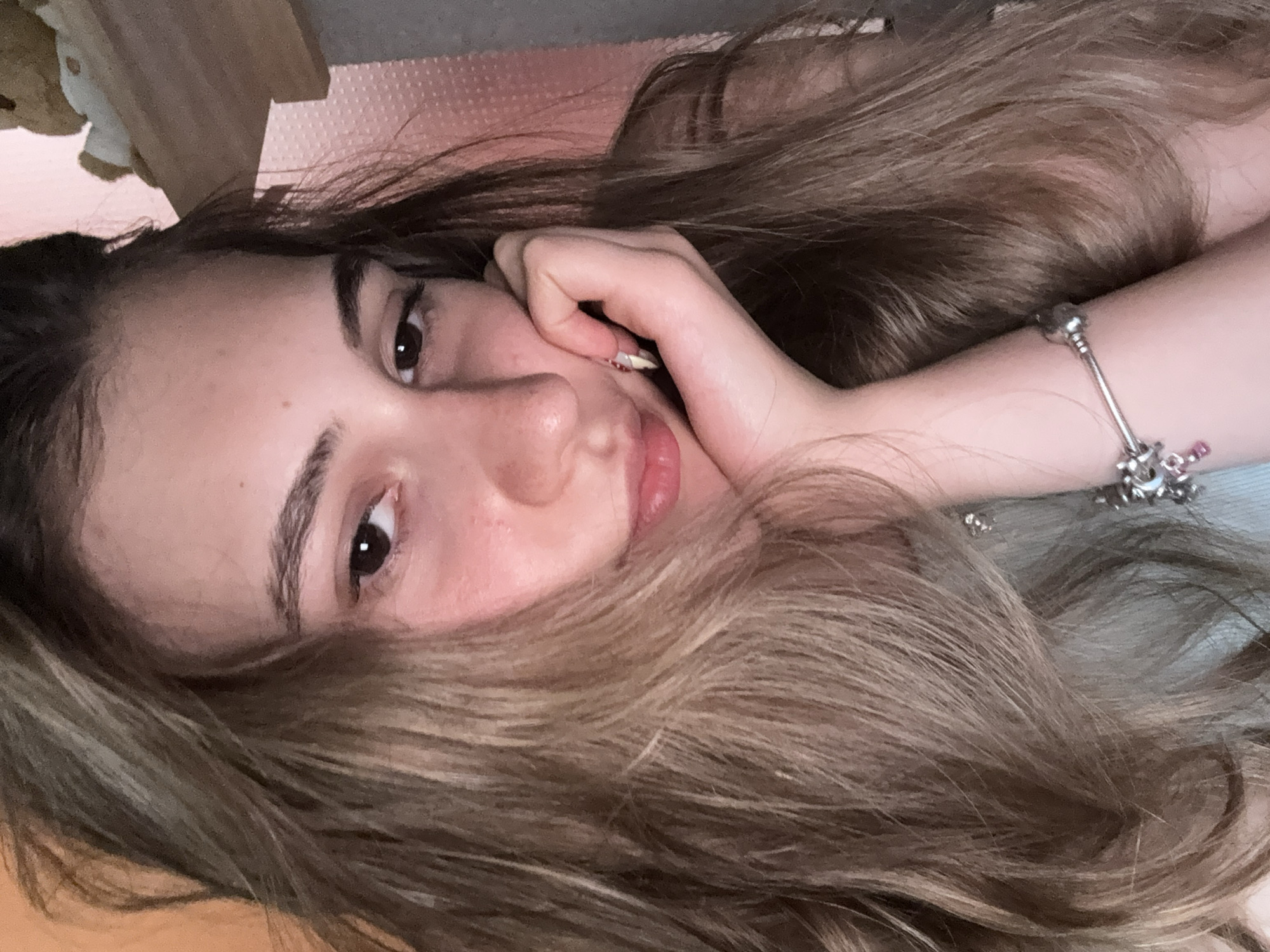 This is a close-up, horizontally oriented, selfie-style photograph of a young person, likely a female, with long, wavy, light brown hair. The subject is lying down, with her head tilted and her hair spread around her face. Her right hand is resting on her cheek, and her left arm is visible, adorned with a silver bracelet featuring charms, including one pink charm.
The subject's facial features are visible: her left eye, eyebrow, nose, and mouth. Her expression appears relaxed and contemplative. The lighting suggests it might be indoors, possibly during the daytime, given the natural light illuminating her face. The background is out of focus, but hints of a light pink textured surface and a wooden object suggest an indoor setting, possibly a bedroom. There is no visible text in the image. The overall mood is intimate and candid.