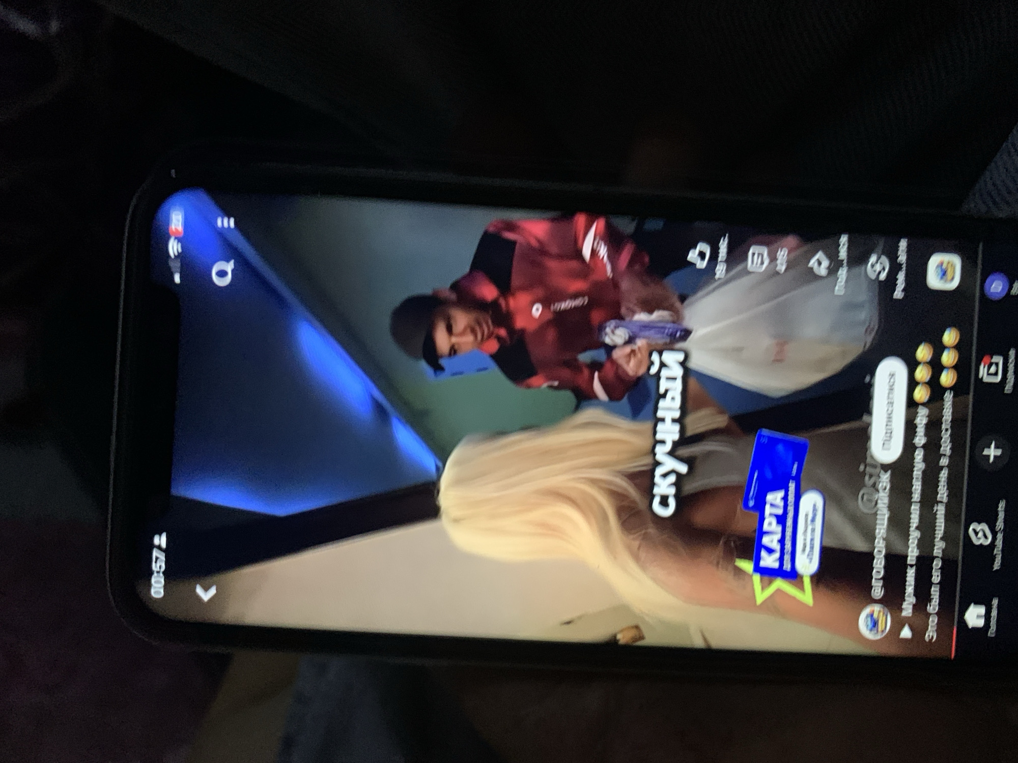 The image is a vertical, slightly blurry, close-up shot of a smartphone screen displaying a TikTok video. The video appears to be a comedic skit or personal story. In the foreground of the video, a woman with long blonde hair is seen from the side, facing away from the camera. In the background, a man wearing a red and black checkered jacket and a dark baseball cap is holding a white plastic bag and a blue item. Text overlaid on the video reads "скучный" (bored) in large white letters, and below that, "Мужик проучил наглую фуру" (The man taught the impudent truck a lesson) and "Это был его лучший день в доставке" (It was his best day on delivery), along with several emoji faces. A blue graphic with the word "КАРТА" (map/card) is also visible. The bottom of the phone screen shows YouTube Shorts interface elements, including icons for Home, Shorts, a plus sign for creation, and a rectangle with a play button. The overall environment within the video appears to be indoors, possibly a street or a delivery area, with some blue lighting effects. The phone itself is held by a person whose hand is partially visible in the foreground, and the background behind the phone is dark and out of focus. No specific details in the image can be used to identify the location as Zhmerynka, Ukraine.