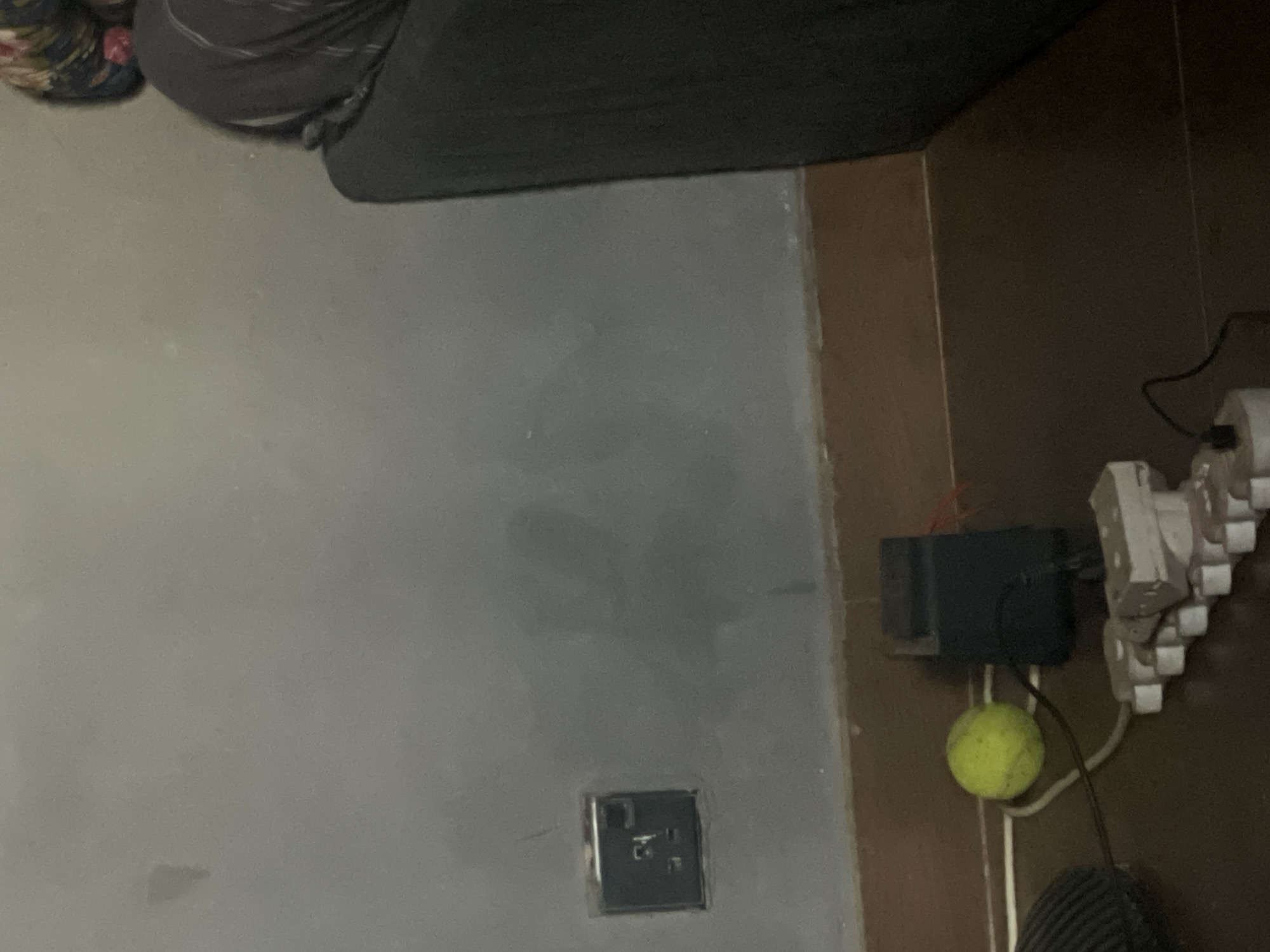 The image depicts a room interior. A portion of a dark sofa is visible in the upper part of the frame, with a person's leg and foot in a patterned slipper on the far left. The wall is painted a dull grey. An electrical outlet is located near the bottom left. Adjacent to the wall is a brown-toned vertical surface, possibly a cabinet or a wall panel. An assortment of electrical equipment and cords are clustered together on the brown surface, with a yellow tennis ball near the equipment. The image was not taken in Kurunduma, Nigeria.