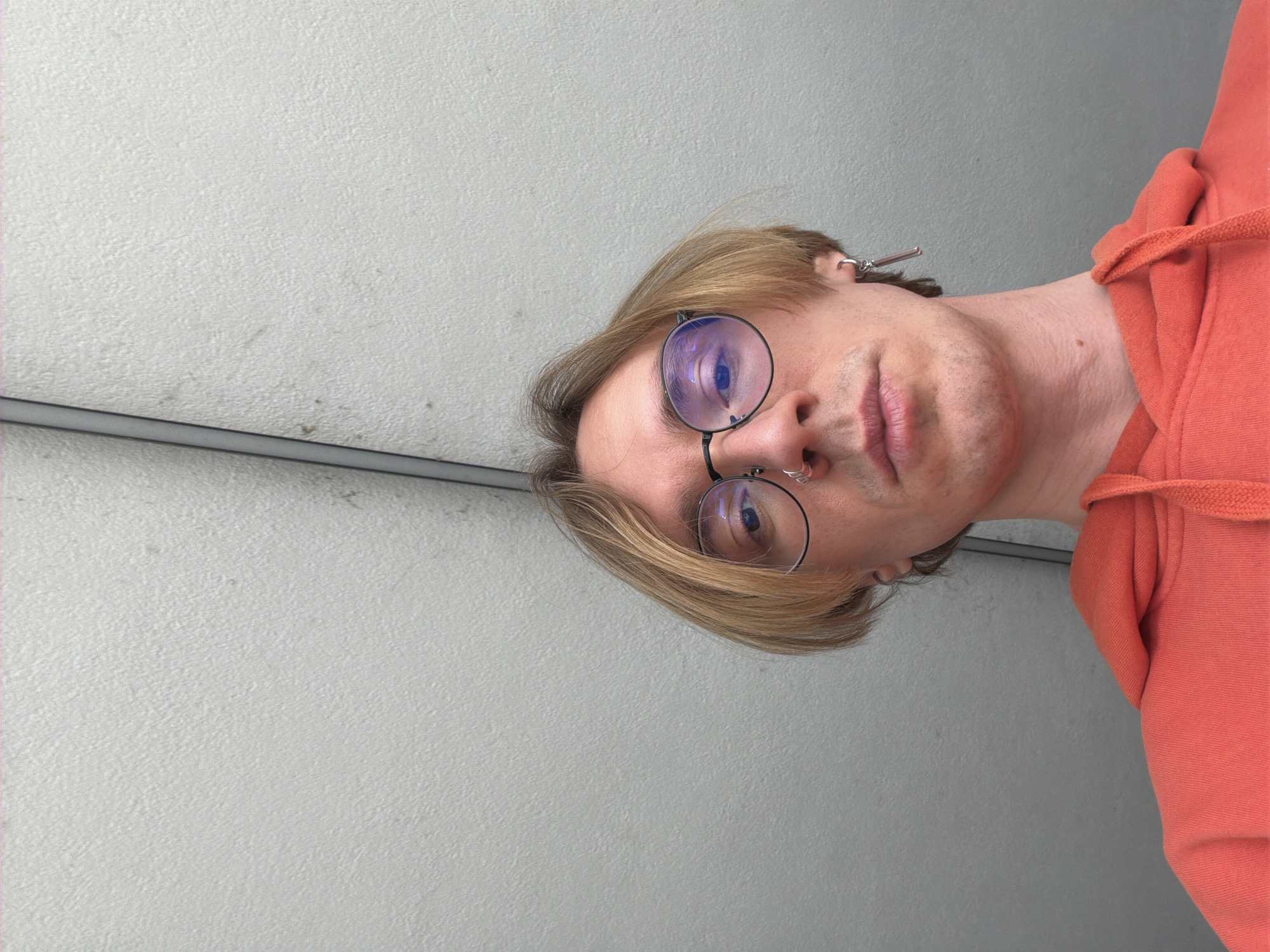 A selfie-style photograph displays a person with blond hair and glasses wearing a coral hoodie. The individual has piercings in their nose and ear. The background is a light grey textured wall. A horizontal line, possibly a seam in the wall, is visible. The photograph appears to have been taken indoors and is at a rotated angle. The location in the city of Budva, Montenegro cannot be ascertained from this image.