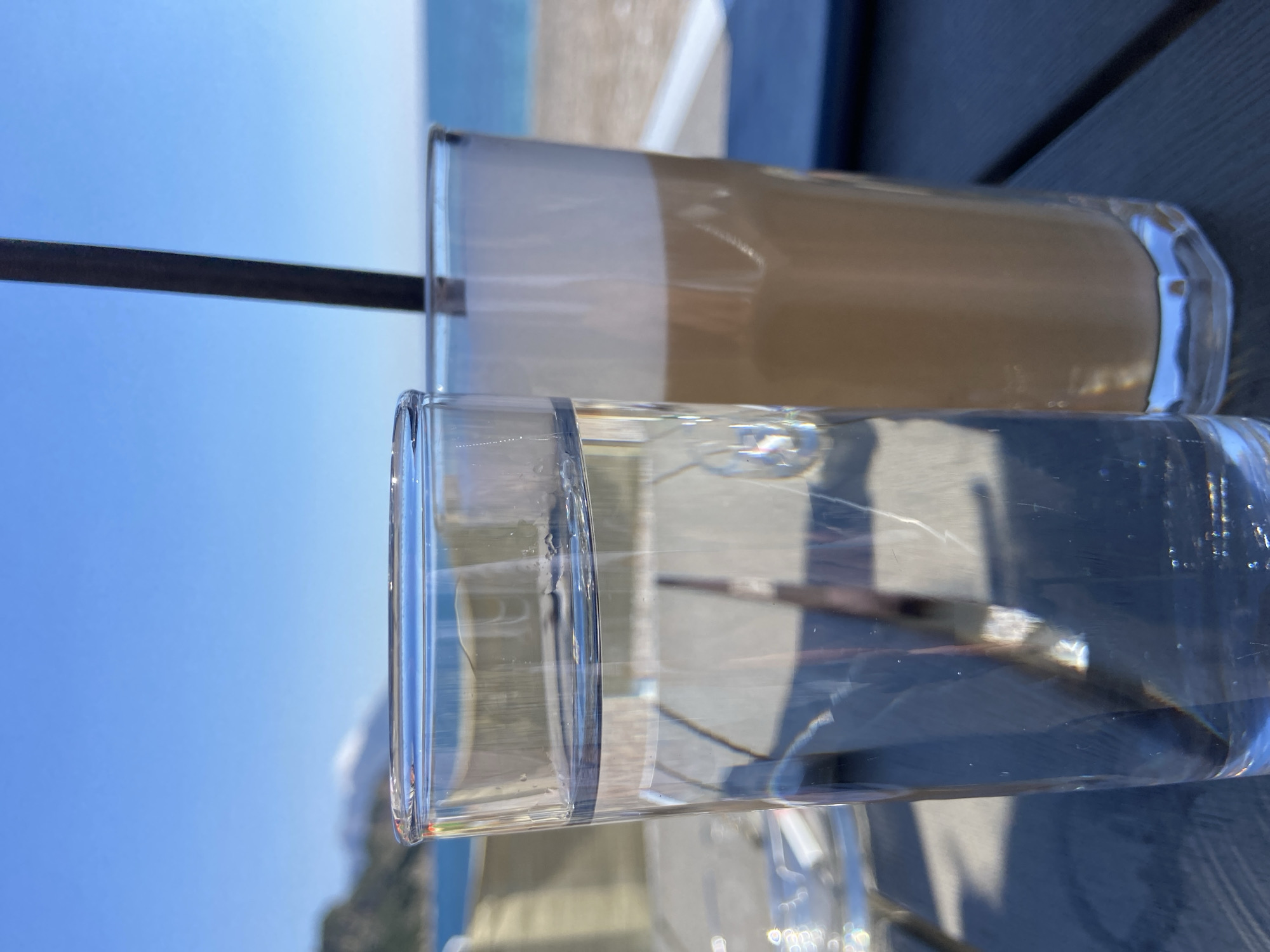 The image depicts a close-up shot of two glasses, one containing water and the other containing a latte-like beverage. The glasses are resting on a dark surface, likely a table, outdoors. The background includes a coastal scene with the sea, a beach, and a mountain or hill. The sky is bright blue. The scene is located in Prijevor, Montenegro.