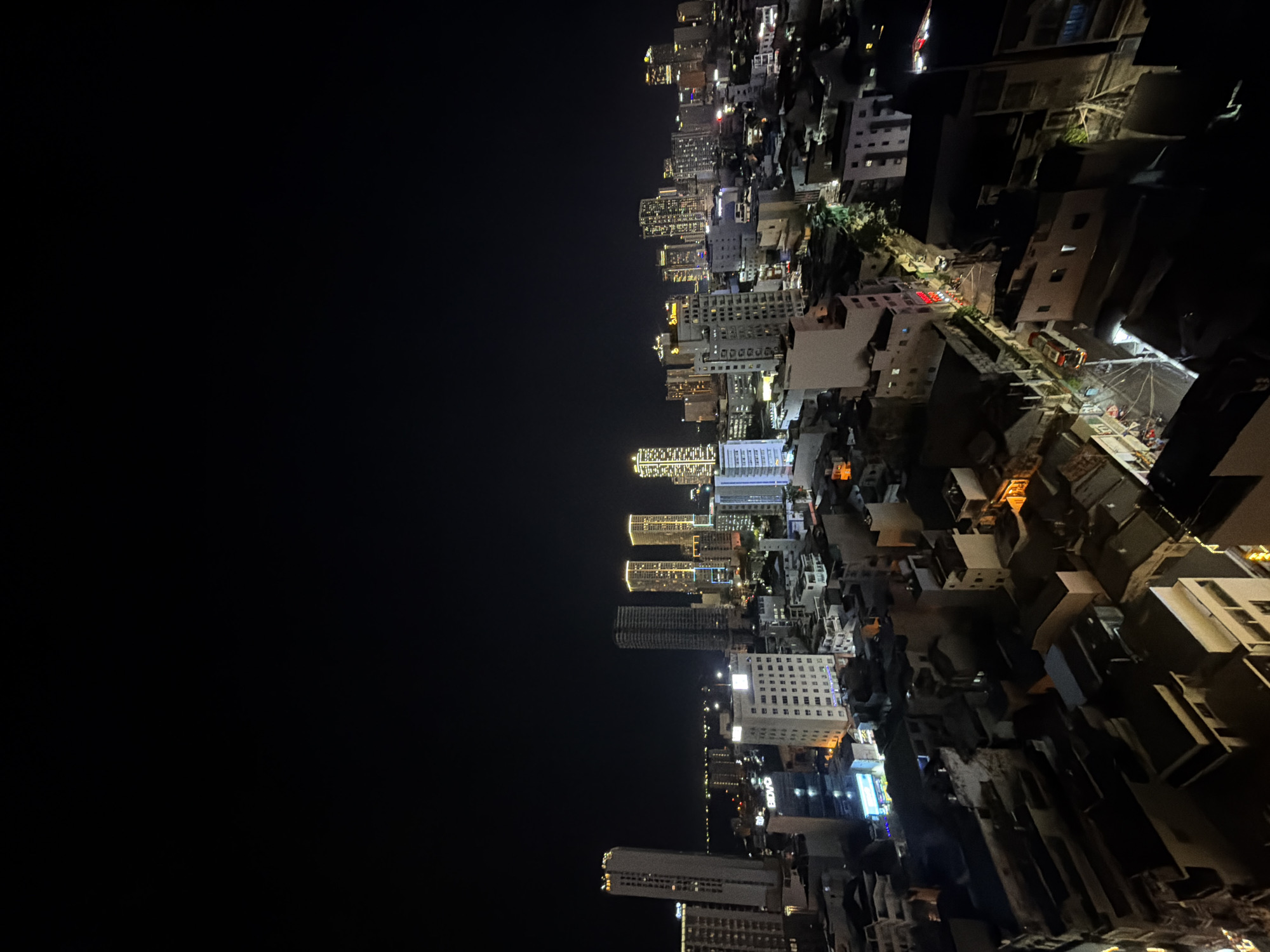 This is an aerial nighttime photograph of a dense urban landscape, likely in Nha Trang Ward, Vietnam, characterized by a mix of modern high-rise buildings and older, lower-rise structures. The sky above is completely black, indicating nighttime.
In the background, a cluster of illuminated skyscrapers dominates the horizon, some with distinctive golden lighting, suggesting they are hotels or prominent office buildings. One taller, slender white building on the far left stands out. The middle ground features a slightly less dense collection of buildings, some also lit, interspersed with darker, less illuminated structures.
The foreground reveals a more tightly packed area of buildings, many of which are darker and appear to be residential or commercial establishments with visible rooftop details and some street-level illumination. A few streets are discernible, with faint lights suggesting vehicular or pedestrian activity. One building has a prominent blue illuminated sign. There is a distinct lack of visible people in the image. The overall impression is a bustling city at night, seen from a high vantage point, with significant artificial light illuminating the urban sprawl.