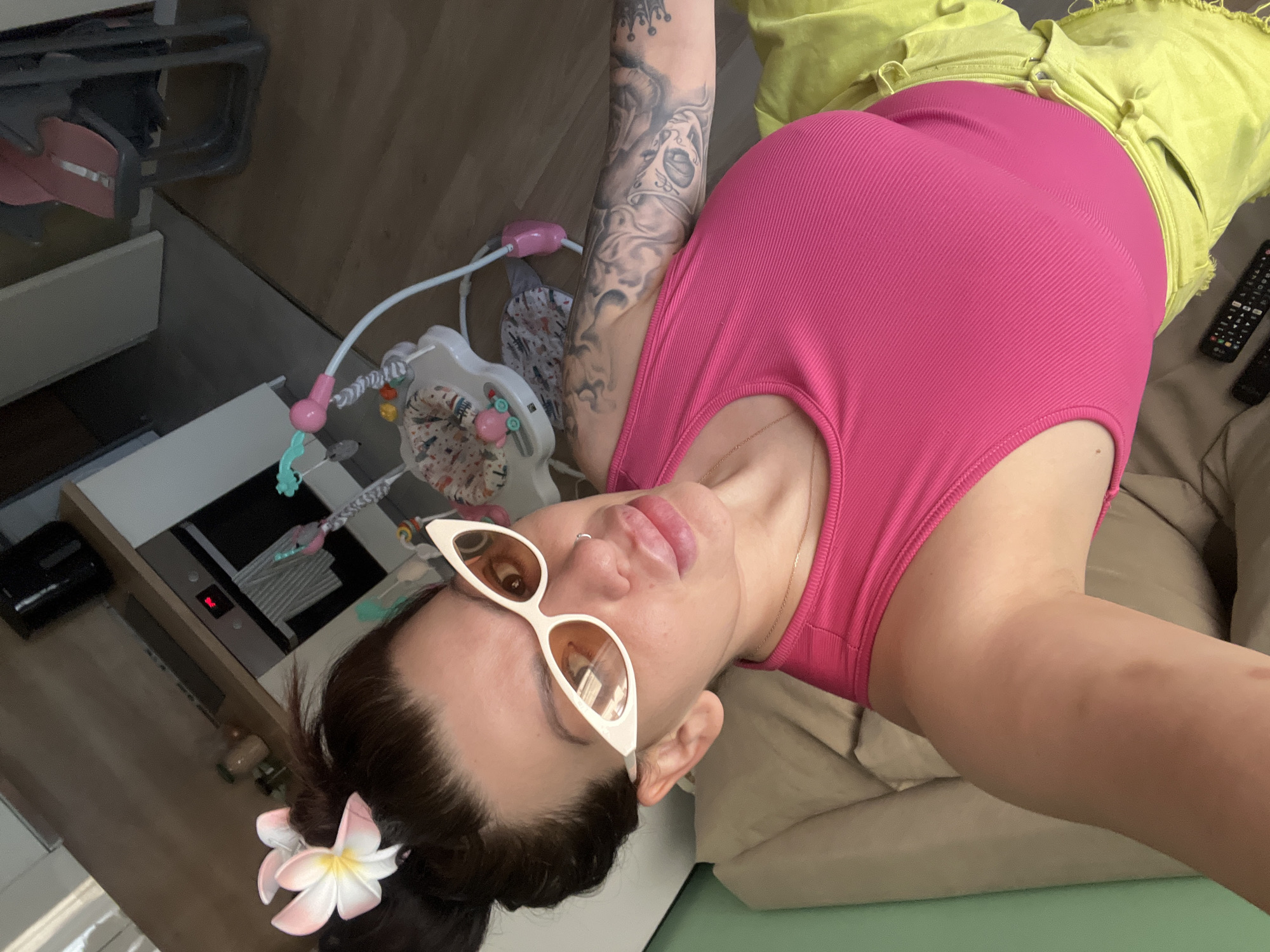 A woman is lying on a bed, facing the camera. She is wearing a pink ribbed tank top and yellow shorts. She has large, plump lips and is wearing white cat-eye sunglasses. A pink and white flower is tucked into her dark brown hair, which is pulled back into a bun. Her left arm, which is visible, is heavily tattooed. In the background, a baby bouncer can be seen. The setting appears to be an indoor living space, possibly an apartment. There is no indication of the location being Plovdiv, Bulgaria.