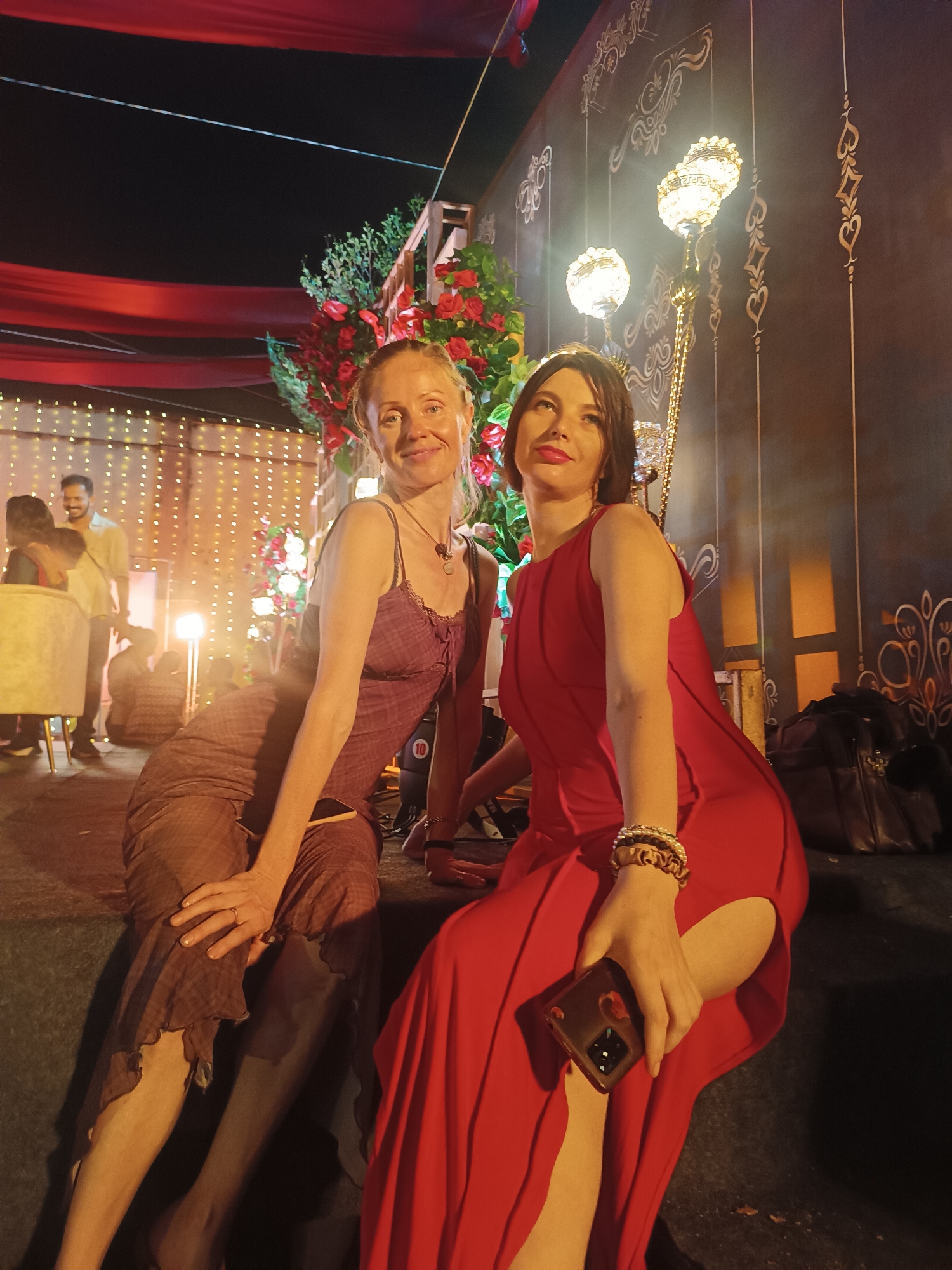 The image features two women seated on a raised platform at what appears to be a festive nighttime event, possibly in Kalamassery, India, given the location hint. The woman on the left has blonde hair, is smiling faintly at the camera, and wears a sleeveless, patterned brown or maroon dress with a ruffled hem and a necklace. A smartphone is visible on her lap.
The woman on the right has dark hair, wears red lipstick, and is looking upwards and to her left with a neutral expression. She is dressed in a sleeveless red maxi dress with a thigh-high slit and has gold-toned bracelets on her left wrist. She holds a smartphone with a decorative case in her right hand.
The background is adorned with elaborate decorations. Red fabric drapes across the top of the scene. Behind the women, large artificial floral arrangements, primarily red roses and green foliage, are prominently displayed. To the left, a wall is covered with warm, glowing string lights, behind which blurred figures of other guests and some furniture can be seen. To the right, a dark wall features intricate gold-colored decorative patterns and two ornate hanging lamps or chandeliers, casting a warm light. A dark handbag rests on the platform next to the woman in red. The overall ambiance suggests a lively and elegant social gathering, likely a wedding reception or a similar celebration, illuminated by artificial light in the evening. There is no clearly visible text in the image.