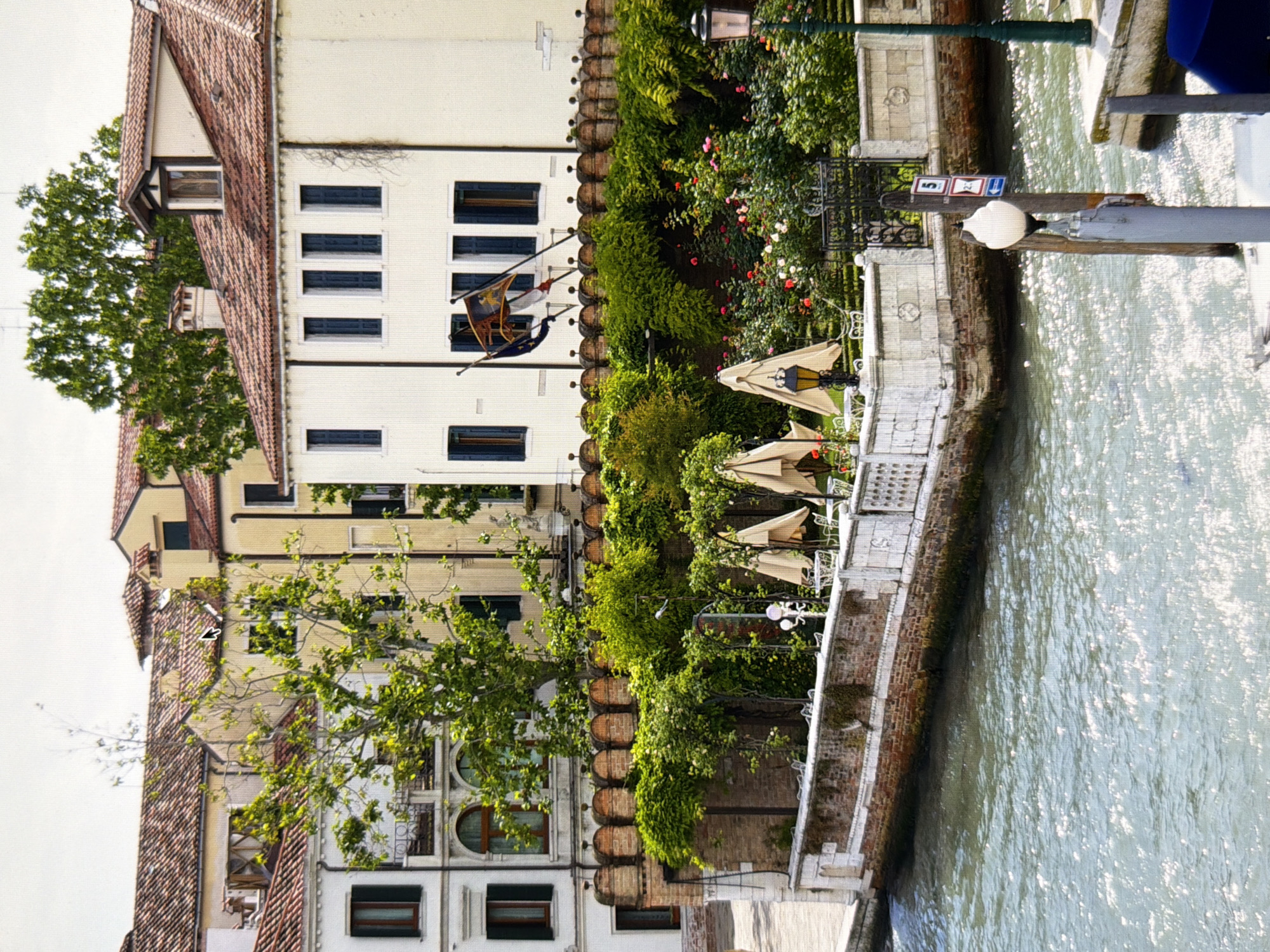 The image depicts a canal-side view of buildings and greenery, with several umbrellas and tables suggesting an outdoor seating area. The architecture appears to be historic, characterized by tiled roofs and cream-colored walls. Trees and lush vegetation are prominent, with flowers blooming in a garden area. Flags are visible flying from one of the buildings. The canal water is rippling, reflecting sunlight. There are no people visible in the scene. The location is identified as being in Kyiv, Ukraine.