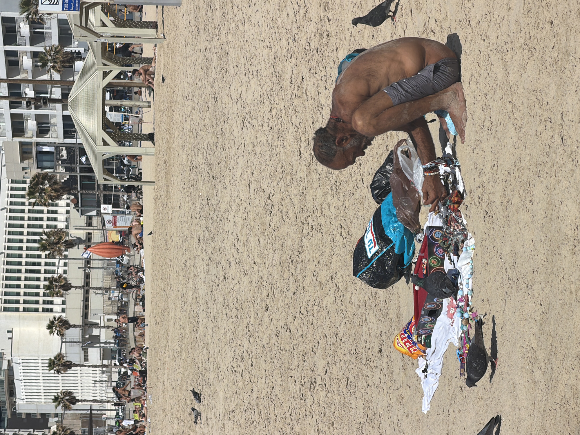 The image shows a beach scene in Tel-Aviv, Israel. A man is bent over, tending to items scattered on the sand next to him. There is a black and blue bag and various objects including pieces of fabric and trash. Two pigeons are near the items. In the background, buildings are visible along the beach, with palm trees and people. There is a structure with a triangular roof. More pigeons are visible around the sand.