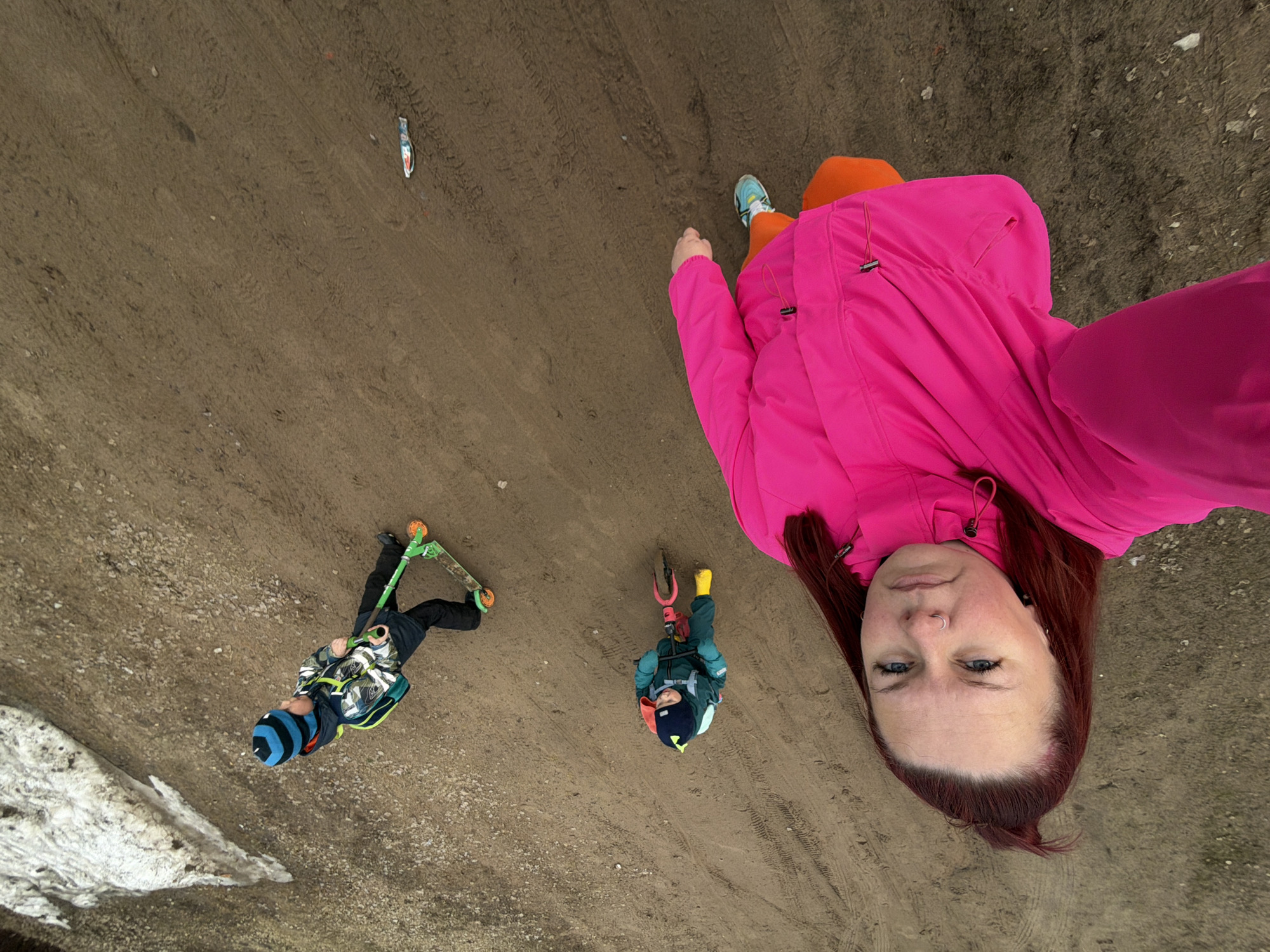 The image shows a woman and two children in an outdoor setting in Chaikovsky, Russia. The ground appears to be dirt or compacted soil with tire tracks visible. A small patch of melting snow is present in the lower left corner. The woman, wearing a bright pink jacket and orange pants, is positioned towards the upper right of the frame. Her face is prominent, with reddish-brown hair. Two children are present in the scene. One is riding a green scooter and wearing a blue and black striped hat with a patterned jacket, and is positioned toward the bottom left of the image. The other child, located closer to the center, is riding a smaller scooter and wearing a blue and yellow hat with a blue jacket. A discarded or dropped container is lying on the ground between the woman and the two children.