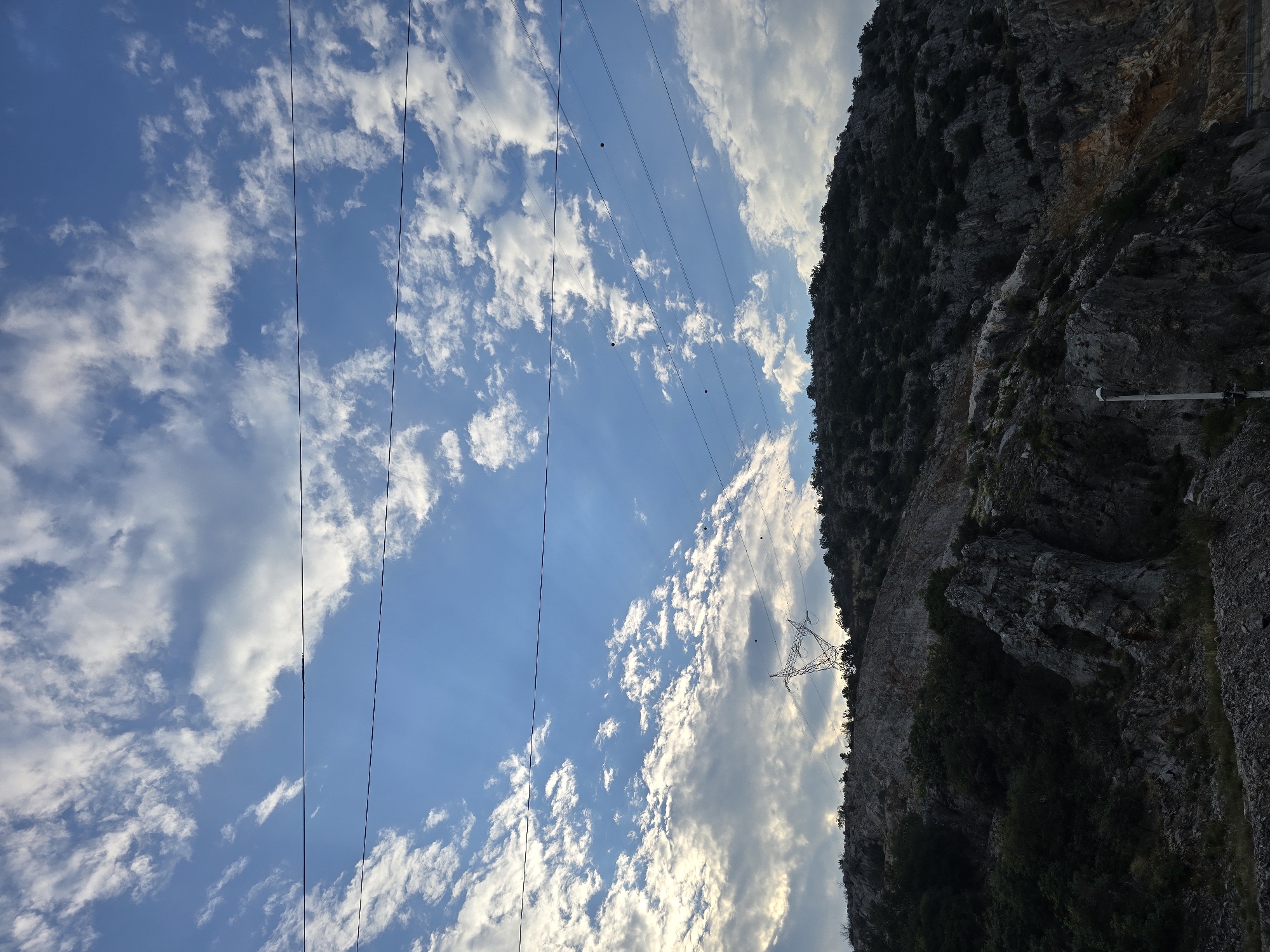 The image depicts a rocky, mountainous terrain with sparse vegetation under a partly cloudy sky. Several power lines stretch across the frame, silhouetted against the blue sky and scattered white clouds. In the mid-ground, a large transmission tower stands on the crest of the hill. The lower portion of the image shows a steep, rocky slope with trees and shrubs growing in crevices. A single street lamp is visible on the right side of the frame, standing near the base of the hill. The scene appears to be an undeveloped or industrial area on the outskirts of Podgorica, Montenegro, possibly near a quarry or construction site. There are no people or animals visible in the image. The lighting suggests it is either early morning or late afternoon.