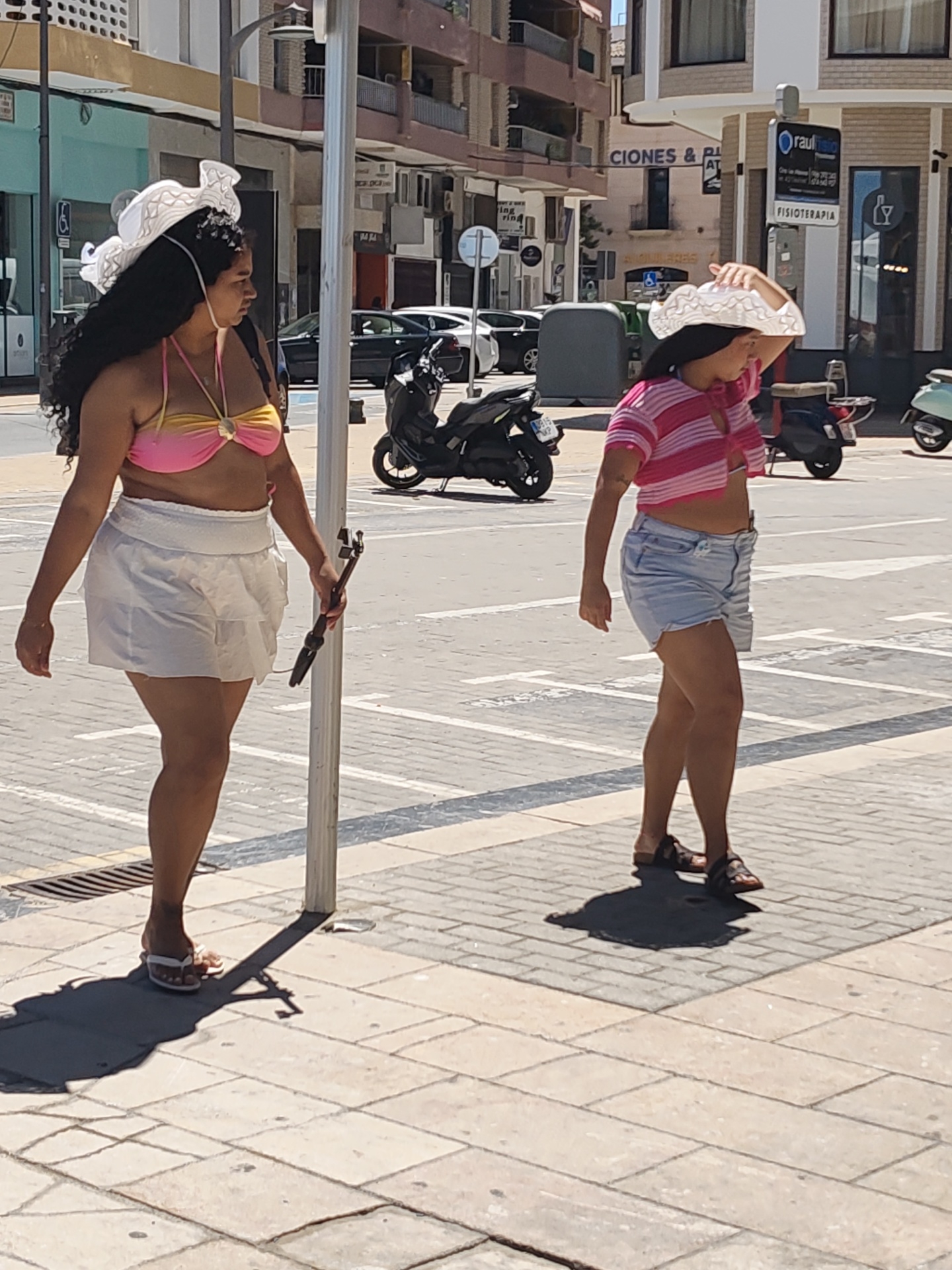 Two women are walking on a paved sidewalk in Dénia, Spain. The woman on the left wears a pink and gold bikini top, a white mini skirt, and white flip-flops. She also has a large white hat and is holding a black selfie stick. The woman on the right wears a pink and white striped crop top, blue denim shorts, and brown sandals. She is also wearing a large white hat and has her right hand holding it to her head. They are both walking away from the camera. In the background, there is a busy street with parked cars and motorbikes, as well as buildings with shops and apartments. A blue sign for "Raul Rubio FISIOTERAPIA" is visible.