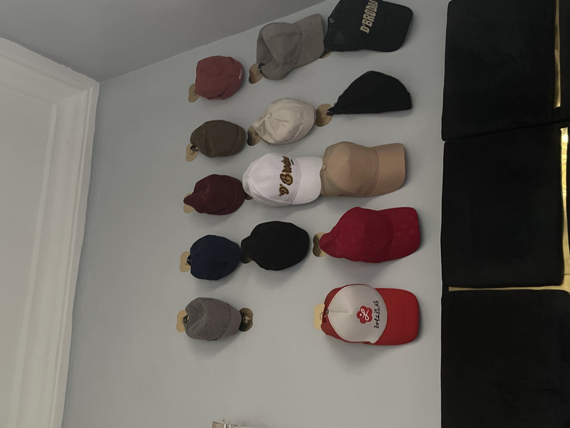 The image shows a collection of baseball caps mounted on a light gray wall. The caps are arranged in two columns and attached to the wall with small, round wooden pegs. The caps vary in color and style. In the background, there is a set of black cushioned panels with gold vertical stripes. The image does not provide any information to suggest a location within Kubwa, Nigeria.