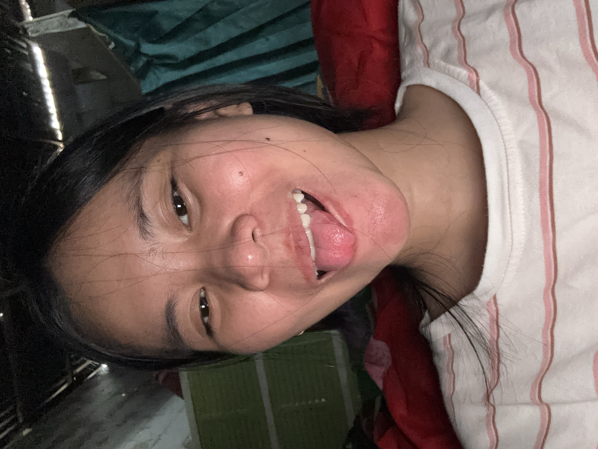 The image is a close-up, vertically oriented portrait of a young person lying down. The subject has dark hair and is looking towards the camera with their mouth open, sticking out their tongue. Their eyes are open, and they appear to be smiling or making a playful expression. The background is dark and somewhat blurred, with hints of green and red fabric, possibly bedding or curtains. There are also some indistinct shapes and a bright, reflective vertical line that could be from artificial lighting or an object in the environment. The subject is wearing a white t-shirt with thin pink and brown vertical stripes. The lighting suggests an indoor setting, possibly in the evening or night due to the darkness. The scene conveys a sense of casualness and playfulness. No text is visible in the image.