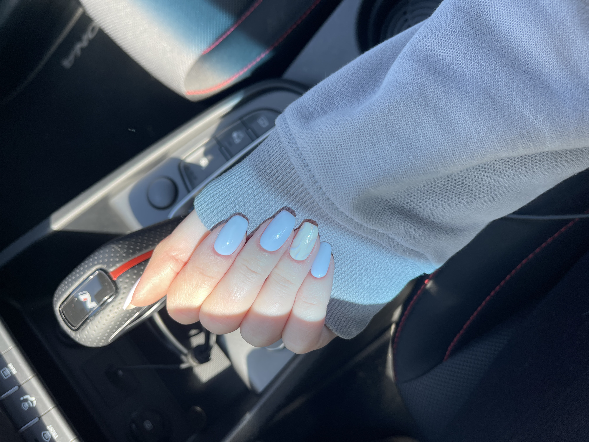 A person is seated in a car, their left hand resting on the gear shift. The person is wearing a light gray, long-sleeved sweater. The person's fingernails are painted a light blue color, except for the ring finger, which is a combination of cream and white. The car's interior includes a black gear shift with red accents, and buttons on the dashboard are partially visible. The car seat is black with red stitching. The location cannot be determined with the given information.