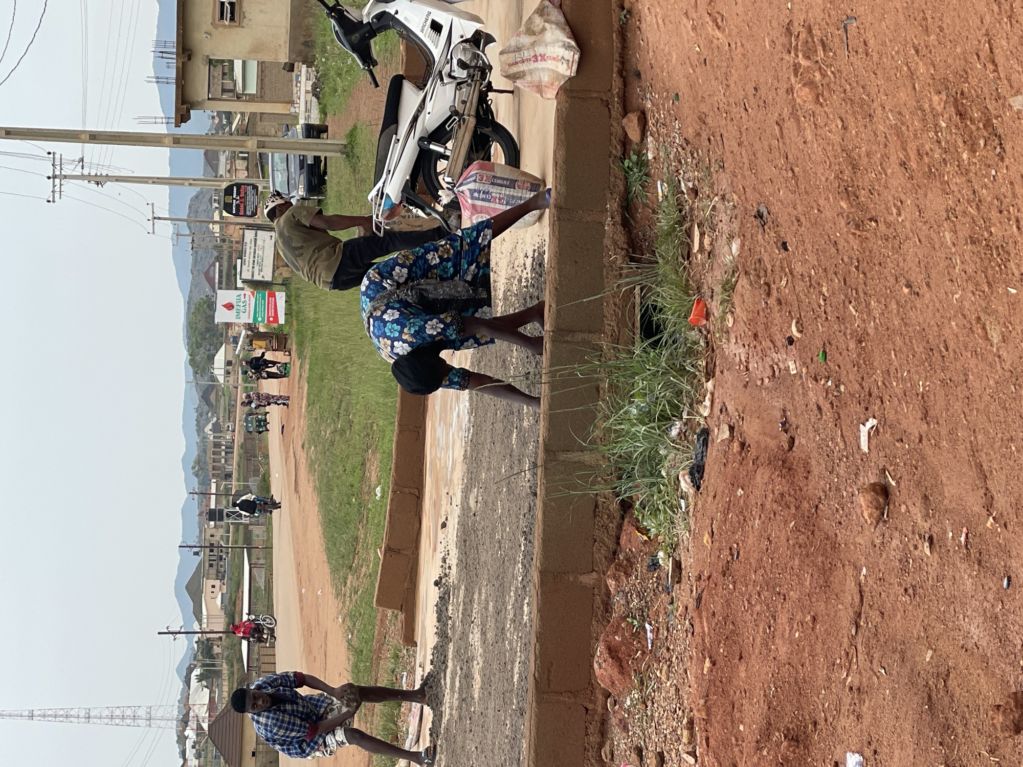 In Jos, Nigeria, on an overcast day, a group of people are engaged in manual labor by the roadside. In the foreground, a young man wearing a plaid shirt and shorts is holding a rock, appearing to be involved in construction or road work. To his right, a woman in a floral dress and headscarf bends down, possibly handling materials. Behind them, a man in a green shirt works near a white motorcycle, with a bag of what appears to be cement nearby. The scene is characterized by dusty, red earth, rough terrain, and a partly constructed road or sidewalk. In the background, various buildings and a tall communication tower are visible, suggesting a developing urban or semi-urban environment. Several vehicles, including motorcycles and a car, are also present on the road, and various advertising signs are posted along the street. The overall impression is one of ongoing construction and daily life in a bustling Nigerian town.
