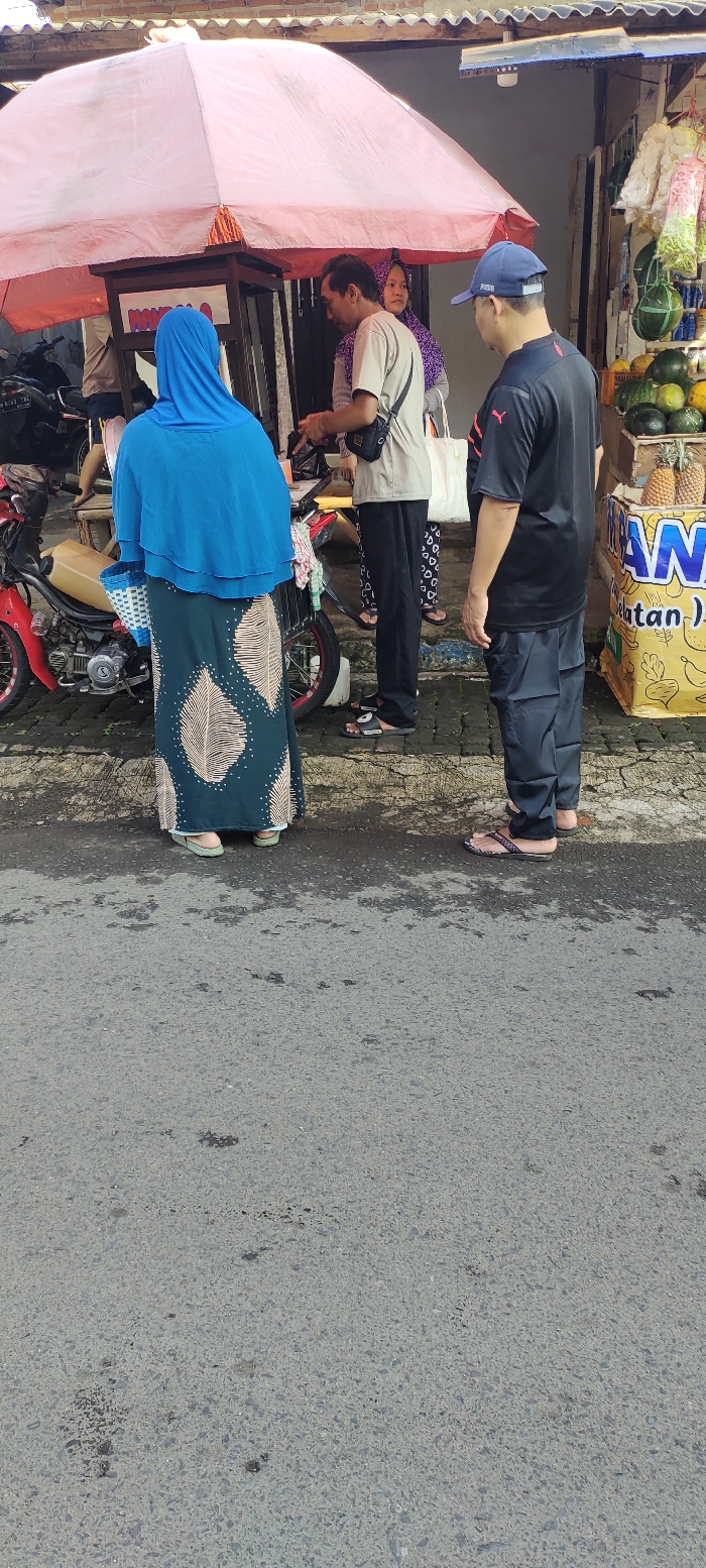A street scene in Kecamatan Lawang, Indonesia (latitude: -7.847, longitude: 112.7), captured from a low angle. Four individuals stand on a paved street, three are facing towards a small food stand, one is standing slightly behind the group, facing away from the stand. The food stand is beneath a pink umbrella, with various items and products displayed. The first individual in line is wearing a light blue hijab and green and white dress, standing in front of the stand. The second individual in line is a man wearing a grey t-shirt, dark pants, and carrying a black bag across his body. He is looking down at the stand. Behind the second individual is a woman with long, black hair wearing a bright pink and purple scarf. She is facing the stand. The last individual in line is a man wearing a black shirt and black pants. His head is turned and facing the camera. All four individuals are standing on a gray pavement, which extends out from the stand. The background is slightly out of focus, but shows a wall with a visible sign with text that reads "AWAN klatan").