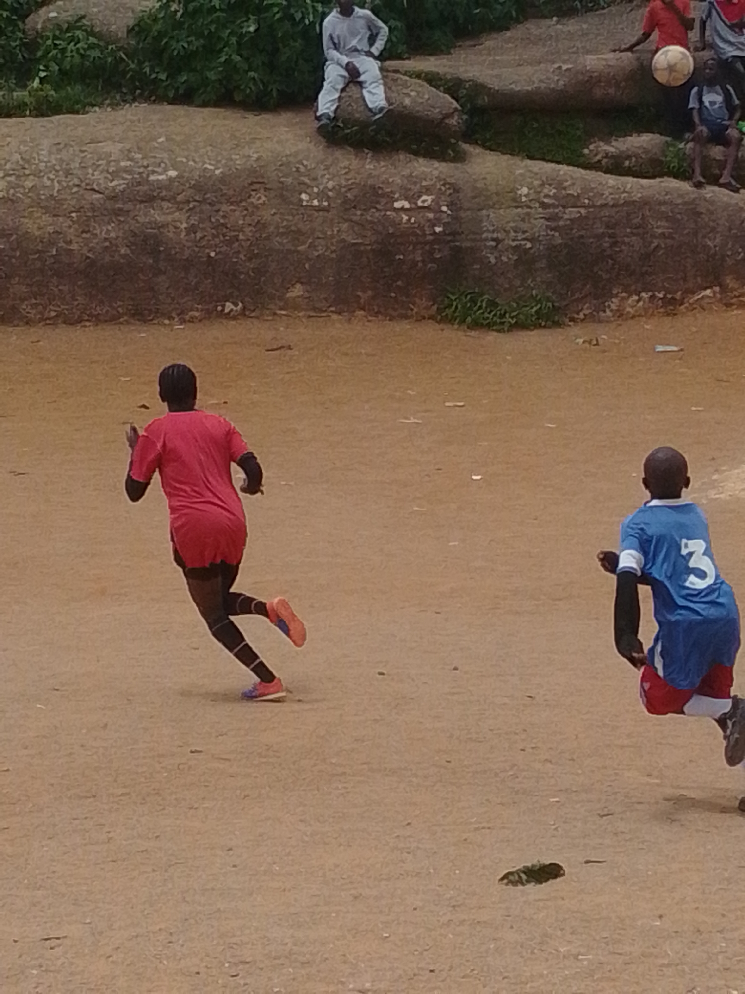 This image depicts an outdoor scene on a dirt field, likely in Jos, Nigeria, where two young boys are engaged in a soccer match. The boy on the left is wearing a red jersey and black shorts, with black and white striped socks. He is captured mid-stride, running with his back to the camera. The boy on the right is wearing a blue jersey with the number "3" prominently displayed on the back, and red shorts. He is also in motion, facing away from the viewer. The ground is a dusty, reddish-brown earth, scattered with small debris. In the background, a large, rocky outcrop is visible, overgrown with green vegetation, suggesting a somewhat rugged natural environment. Several other figures, including children and adults, are partially visible on top of the rocks, observing the game or resting. One individual in the background is seated on the rocks wearing light-colored clothing, and another, a child, is seated with a soccer ball. The lighting suggests it is daytime, with no clear indication of specific time of day. The overall atmosphere appears to be casual and recreational.