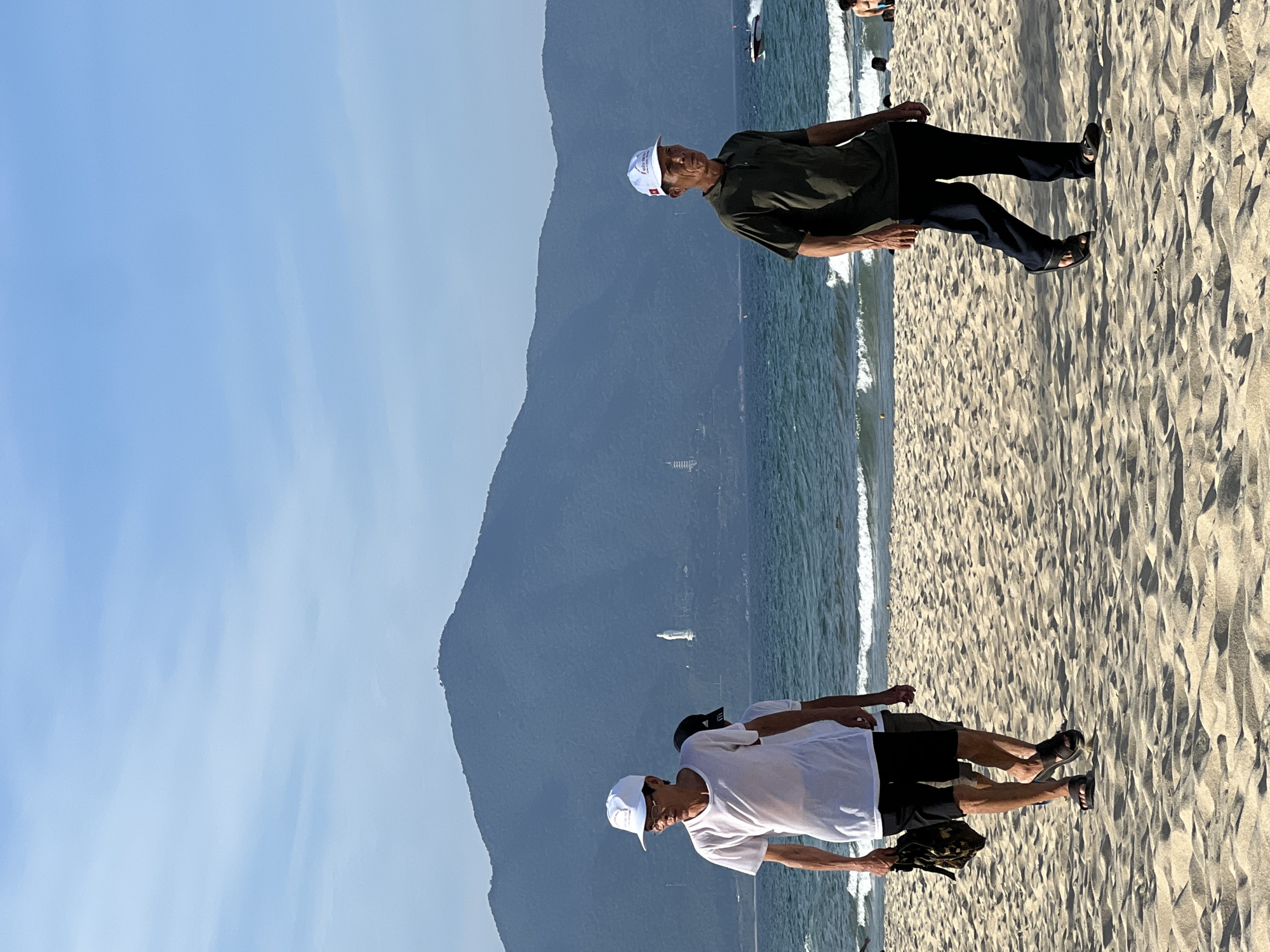 This is a photograph taken at My Khe Beach in Da Nang, Vietnam, during daylight hours. The image features three individuals walking on the sandy beach towards the viewer. In the foreground, a man wearing a white t-shirt, black shorts, and a white baseball cap is walking on the left. He is carrying a dark bag in his left hand and wearing sandals. Behind him, partially visible, is another person. To the right, a man in a dark olive-green short-sleeved shirt, dark pants, and a white baseball cap is walking. He is also wearing sandals. The background displays the ocean with gentle waves and a distant mountain range. On the slopes of the mountain, a tall white statue and several smaller structures are visible. A speed boat can be seen in the water to the right, and other beachgoers are present in the distance. The sky is clear blue with some light cloud formations.
