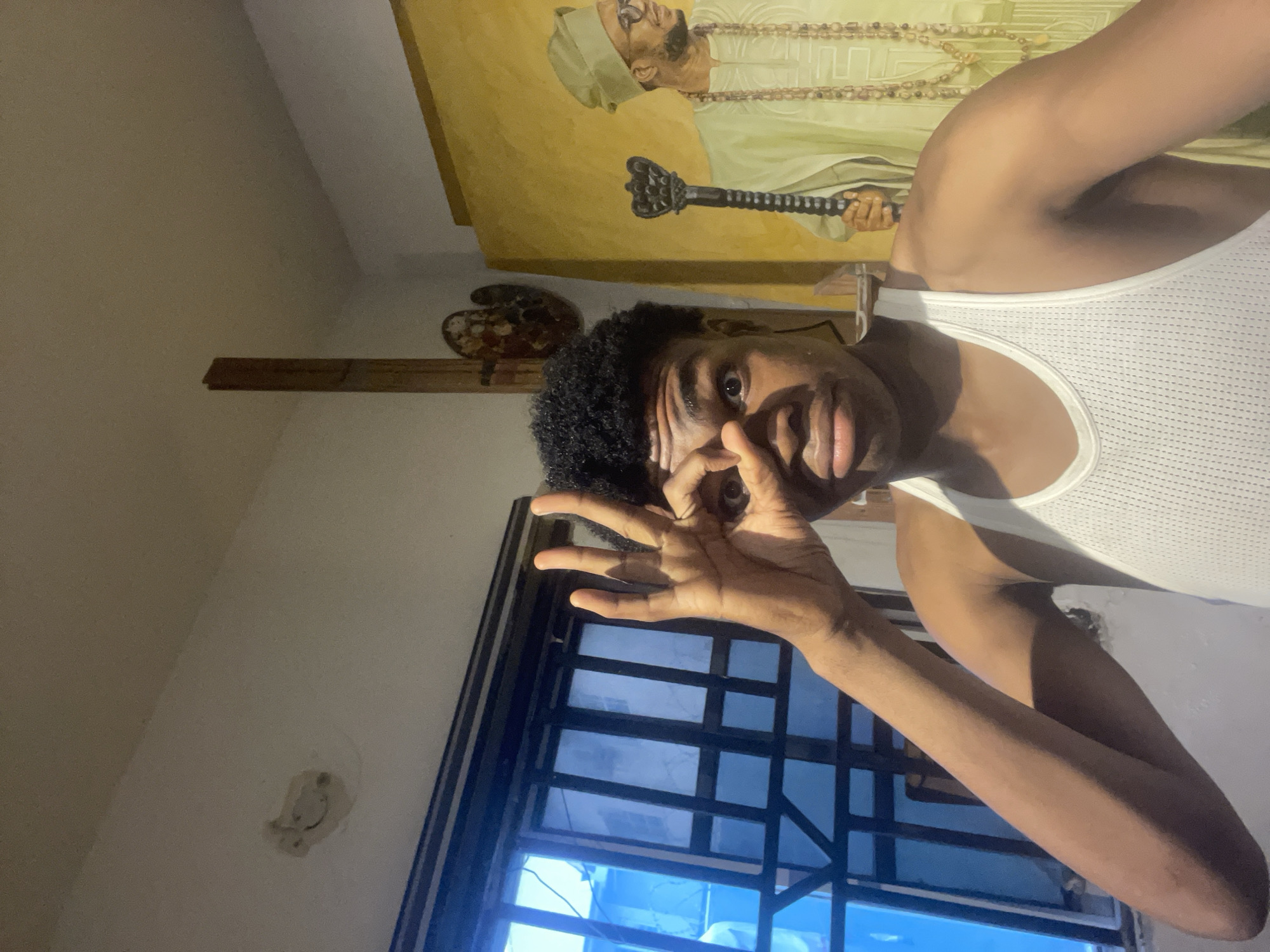 A young Black man with curly hair, wearing a white tank top, is shown in a selfie. He is making a gesture with his right hand, forming a circle with his thumb and index finger, and holding it up to his eye. His left hand is raised slightly. In the background, there is a painting of a man wearing traditional Nigerian attire and a cap, holding a ceremonial staff. To the left, a window with bars and blue-tinted glass is visible. The setting appears to be an indoor space, possibly an art studio or residence, with textured walls and what looks like a wooden easel or stand behind the man. The lighting is warm and casts shadows.