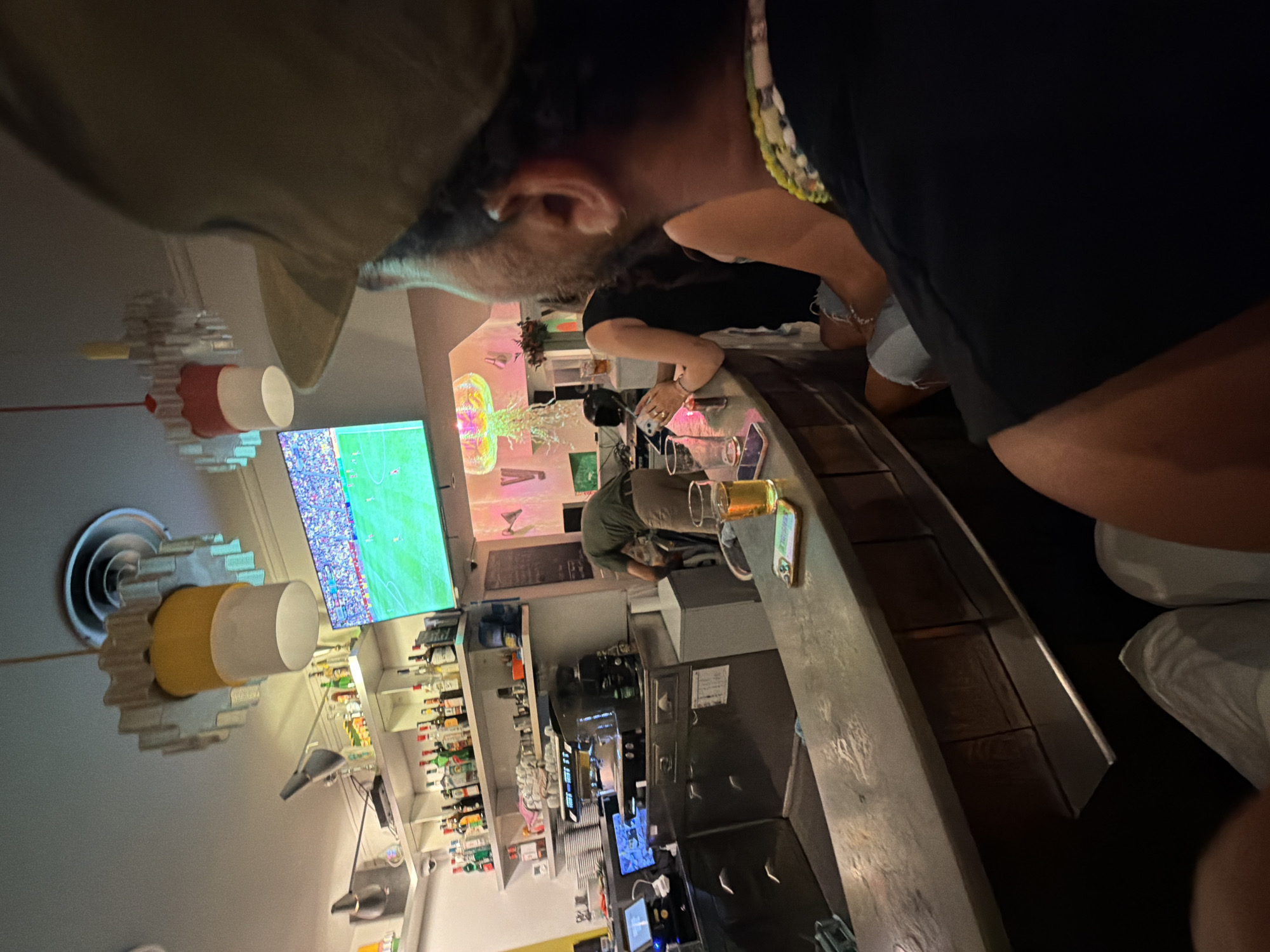 The image depicts a lively bar scene, possibly in Lyon, France, with a prominent flat-screen television displaying a football match in the background. A man wearing a baseball cap is in the foreground, his back to the camera, looking towards the screen. Other patrons are visible at the bar, some engaged in conversation and drinks. A bartender is attending to the counter, and shelves stocked with bottles of alcohol line the wall. The bar area is dimly lit, with ambient lighting from the TV and decorative lamps creating a warm atmosphere. The overall impression is one of a casual and social gathering, enjoying the game and each other's company.