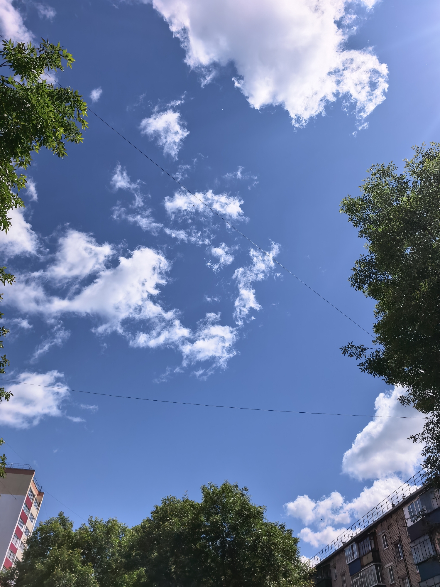 The image shows a clear blue sky with scattered white clouds, taken from a low angle looking upwards. Green trees frame the top left and right of the image, with branches extending into the frame. Two separate buildings are visible at the bottom. On the left, a tall building with white and red vertical stripes is partially shown. On the right, a brick apartment building with balconies runs along the bottom edge. Two thin black wires stretch across the sky, one higher than the other. The scene appears to be an urban environment on a sunny day. The location is the city of Sterlitamak, Russia.