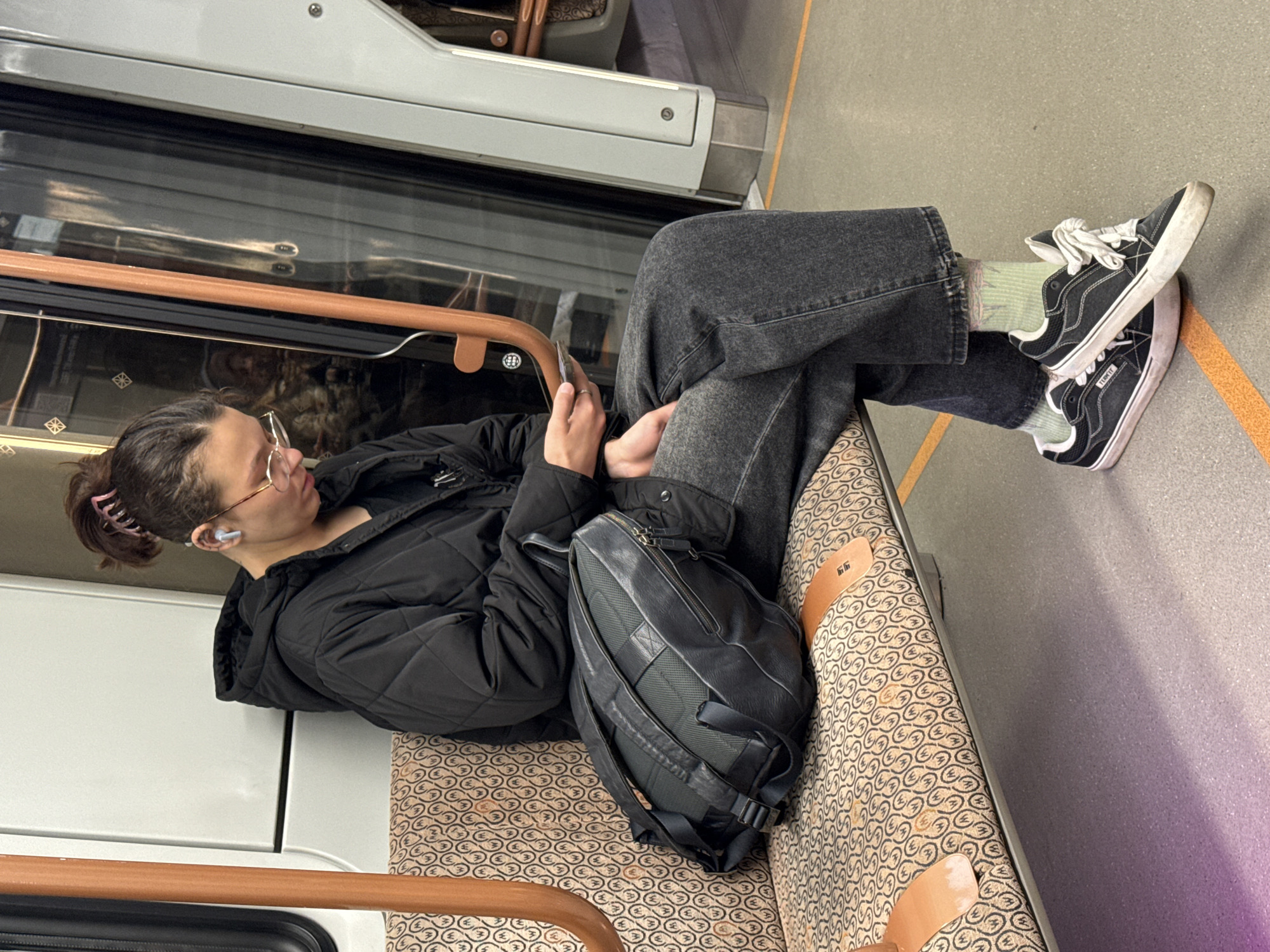 An individual is seated on a subway car with a brown patterned seat, holding a smartphone and looking at it. The subway car's window is to the left of the person. The individual has gray jeans and black tennis shoes stacked on top of each other. A black backpack rests on the seat next to them. The location is likely a subway in the city of Moscow, Russia, as suggested by the subway car's style and design.
