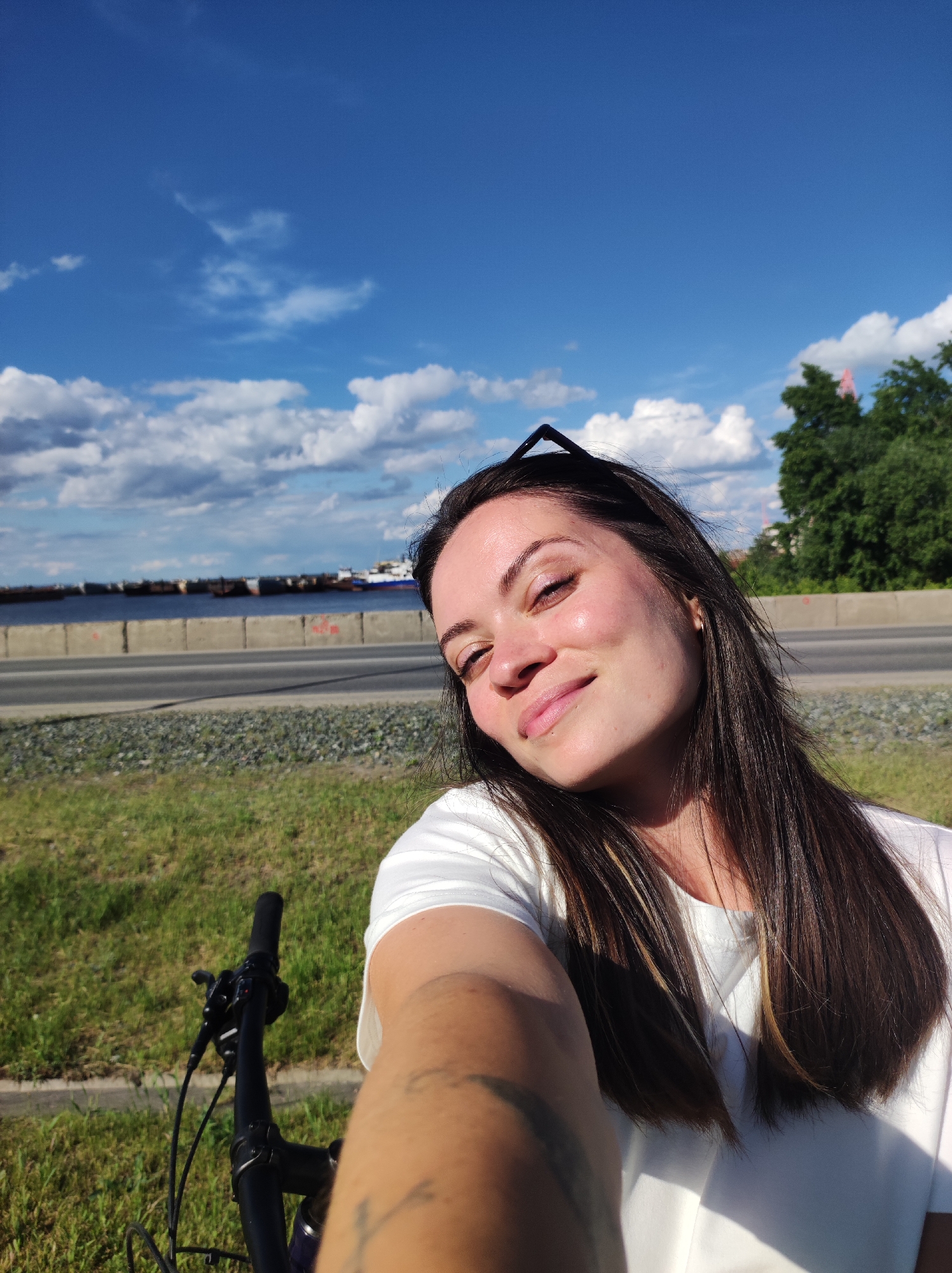 A woman is taking a selfie while riding a bicycle in what appears to be Chaikovsky, Russia. She is fair-skinned with dark hair and is wearing a white t-shirt. Her sunglasses are resting on top of her head. A portion of the bicycle handlebars and her outstretched arm are visible.
In the background, there is a road with a low concrete barrier, followed by a body of water with several ships or barges. Beyond the road, there is a grassy area and some trees. The sky is blue with scattered white clouds.